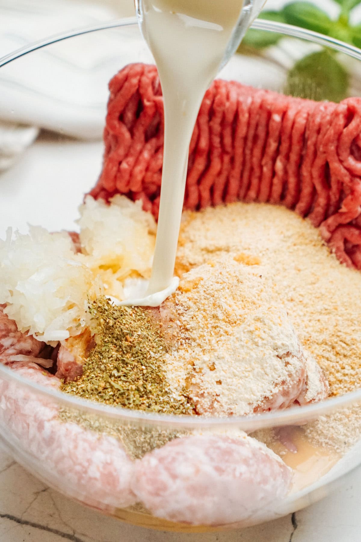 A bowl containing ground meat, breadcrumbs, chopped onions, seasonings, and being combined with a liquid being poured in.