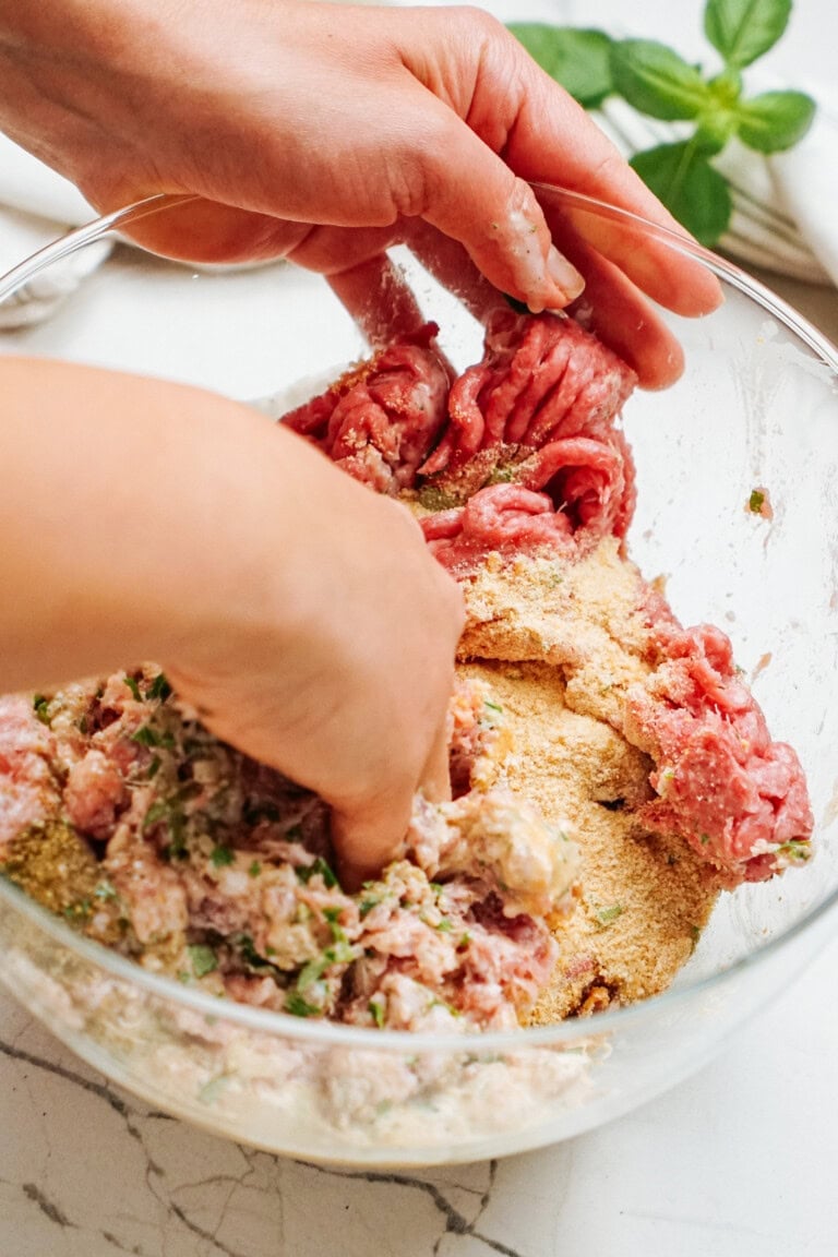 Hands mixing ground meat, breadcrumbs, and seasonings in a glass bowl on a marble countertop.