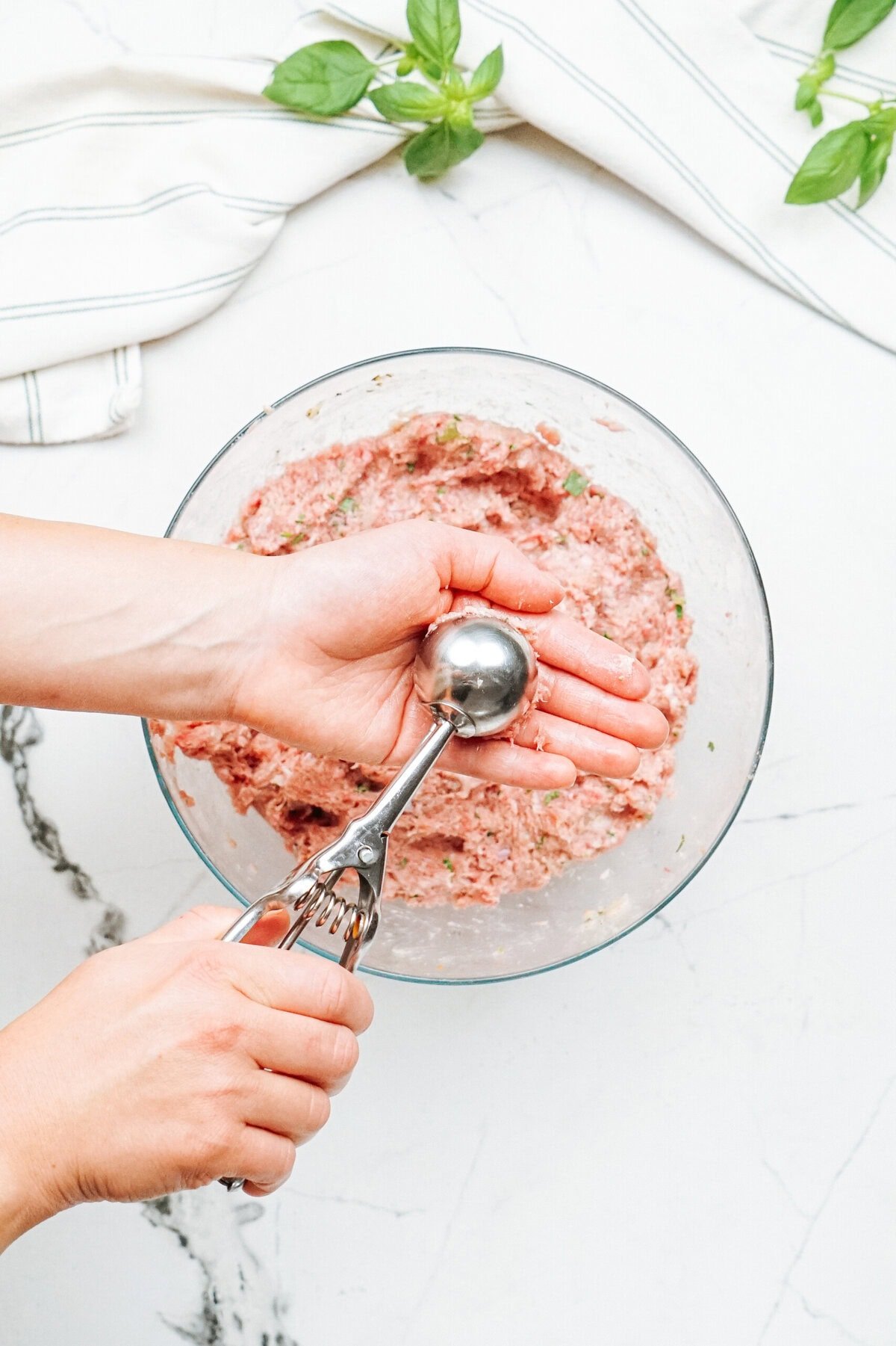 A person uses a metal ice cream scoop to portion meatball mixture from a glass bowl on a white marble countertop with basil leaves and a cloth nearby.