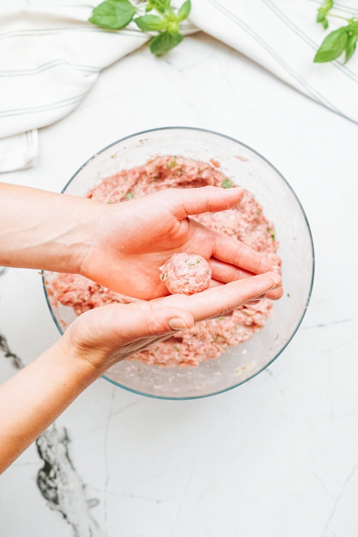 Person forming a meatball with their hands over a bowl of ground meat mixture on a white countertop with herbs in the background.