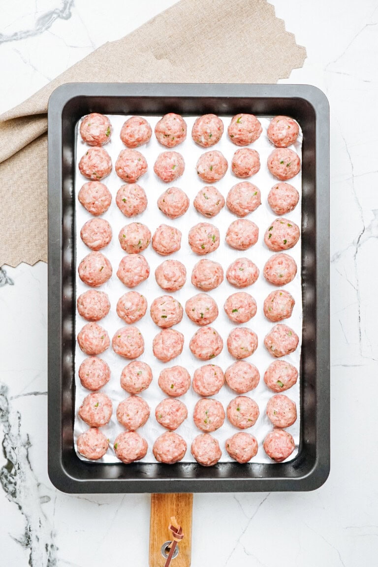 A baking tray filled with evenly placed raw meatballs, ready to be cooked, rests on a marble countertop. A beige cloth is partially visible in the background.