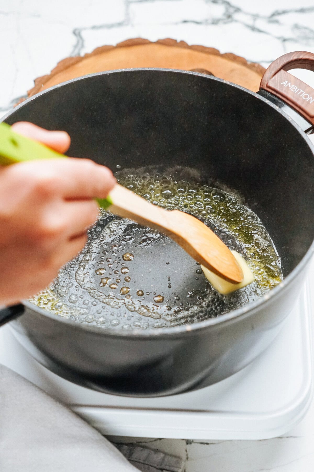 A person melts butter in a black saucepan, stirring with a wooden spoon on a stove.