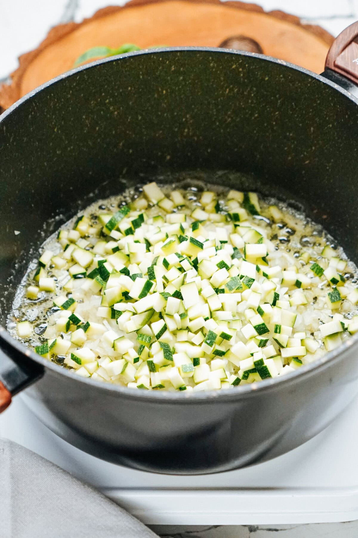 Diced zucchini and onions are sautéing in a black pot on a stovetop.