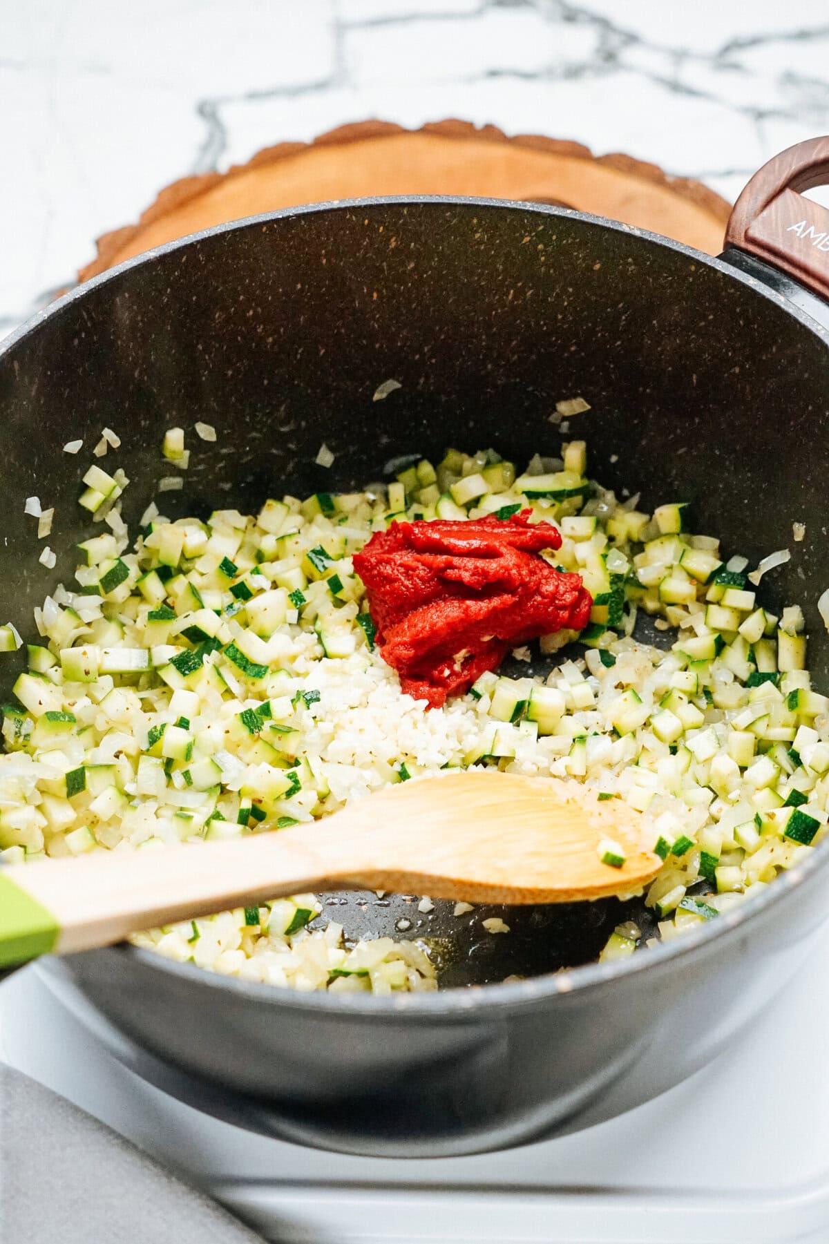 A pot containing chopped zucchini and onions with a dollop of tomato paste on top, being stirred with a wooden spoon.