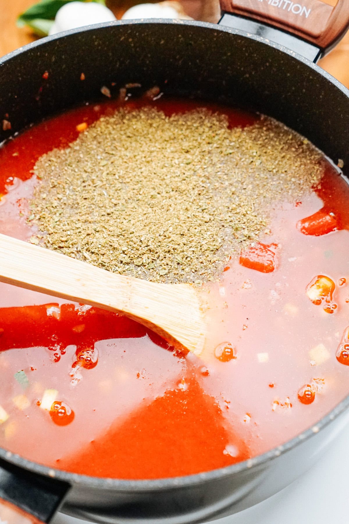 A wooden spoon stirs tomato soup in a black pot, with a layer of dry herbs sprinkled on top.