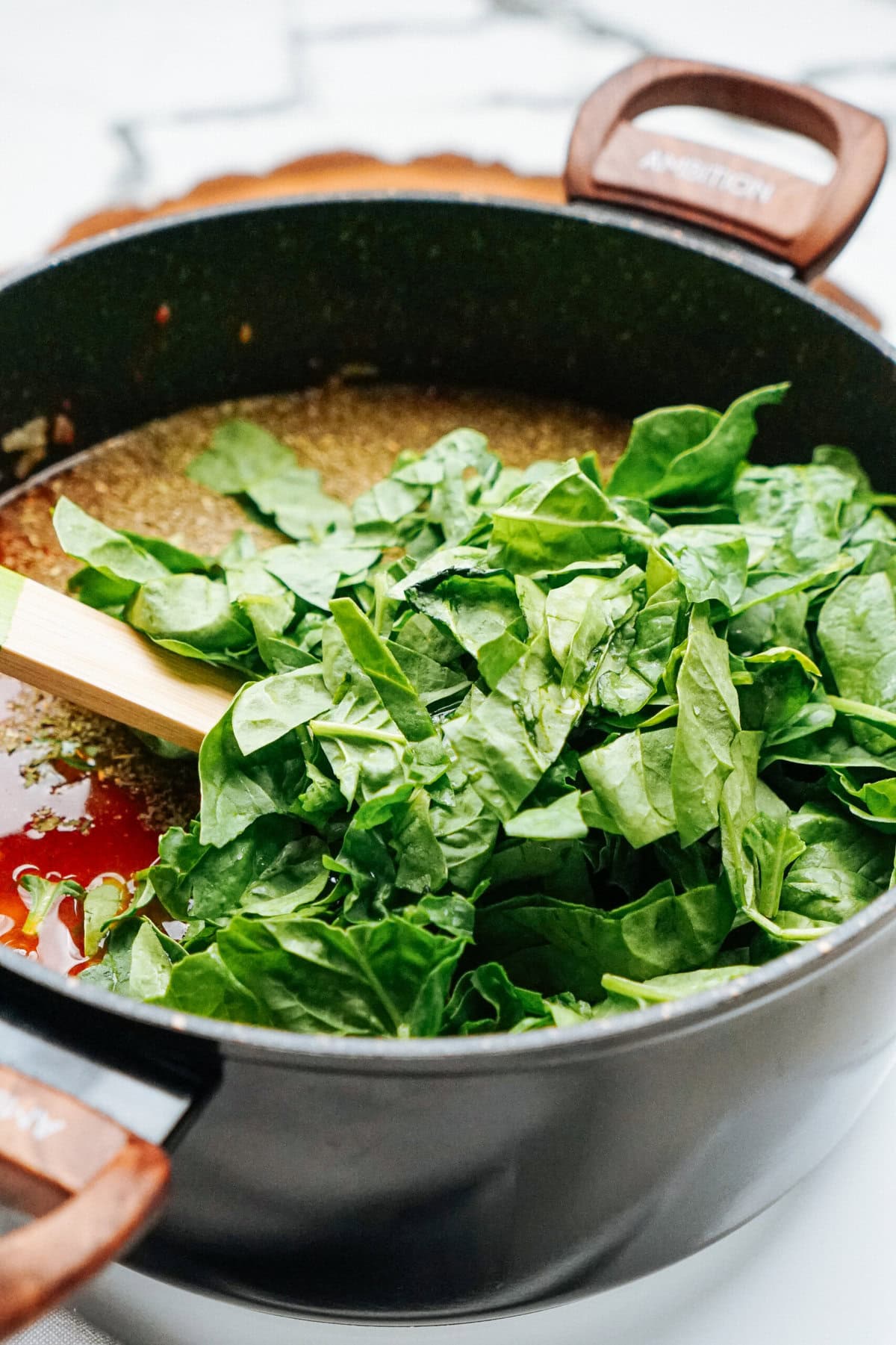 A black pot on the stove contains a mixture of ingredients, including a large amount of fresh spinach. A wooden spoon rests in the pot.