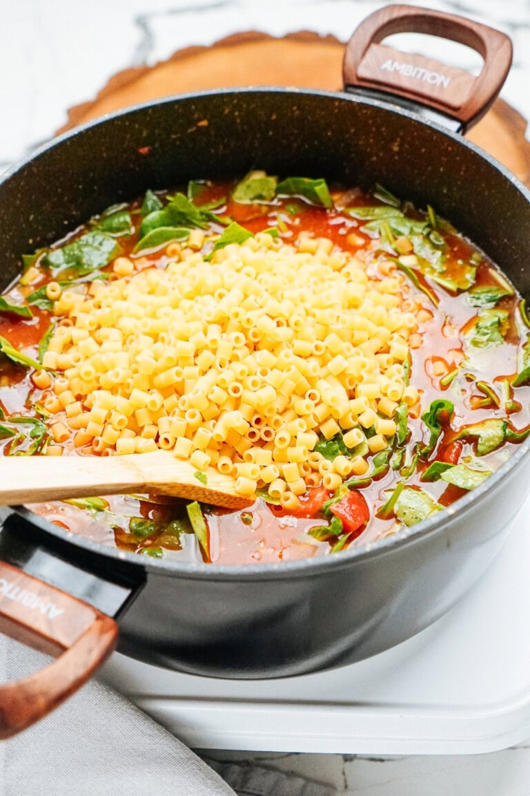 A pot filled with a tomato-based Italian meatball soup containing chopped greens and pasta, with a wooden spoon resting on the edge.