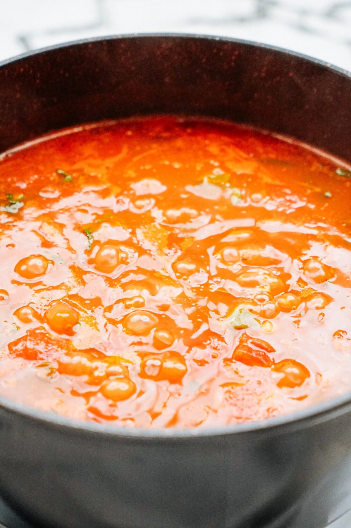 A close-up of a pot filled with bubbling tomato soup or stew, showing a rich red-orange color with visible herbs.