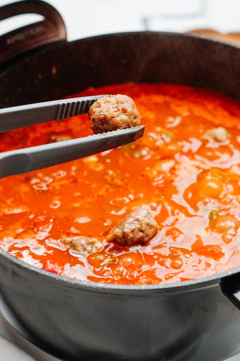 A close-up of meatball soup in a black pot with metal tongs holding a meatball above the soup.