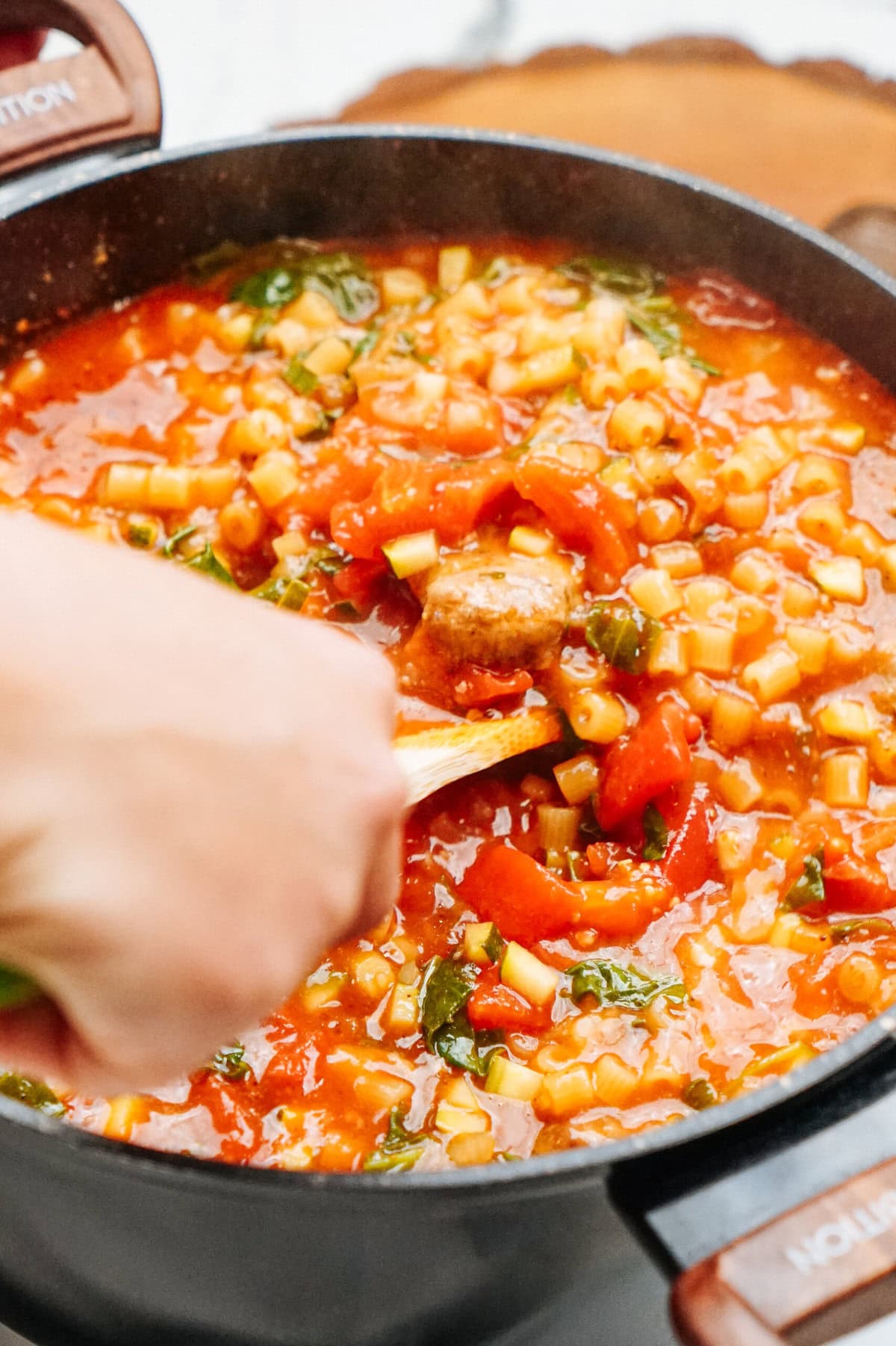 A hand stirring a thick stew of tomatoes, pasta, and vegetables in a black pot.