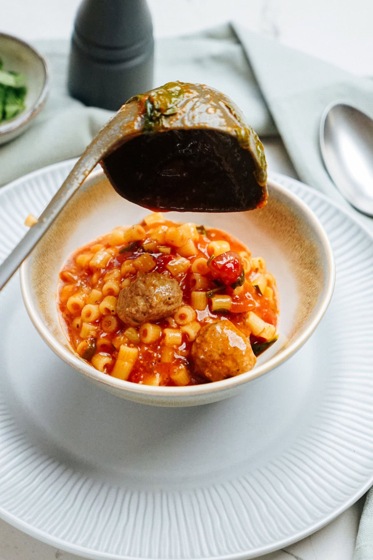 A ladle pours tomato soup with macaroni and meatballs into a white bowl placed on a matching plate.