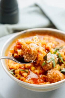 A close-up of a spoonful of soup containing meatballs, pasta, diced vegetables, and herbs with a bowl of the same soup in the background.