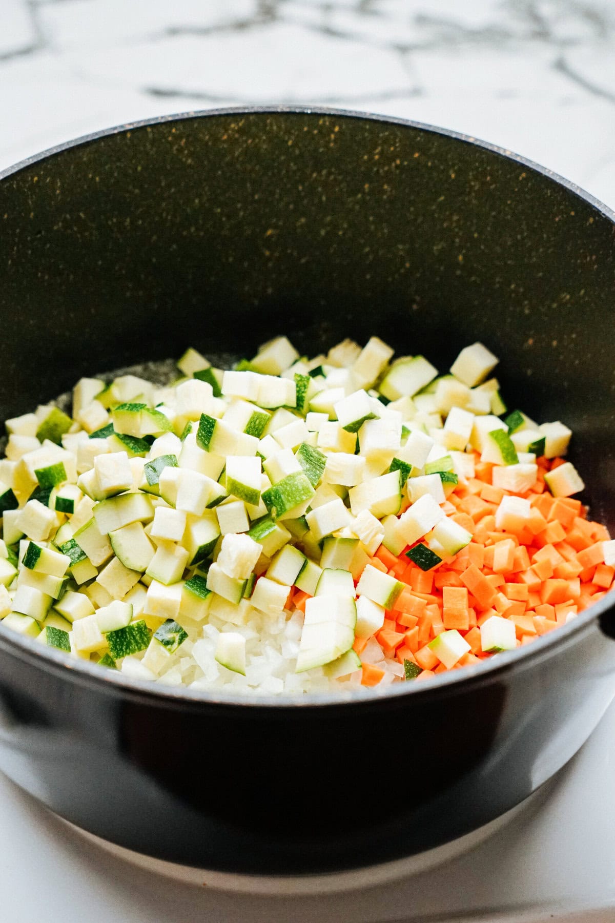 A pot filled with chopped zucchini, carrots, and onions on a blurred kitchen countertop background.