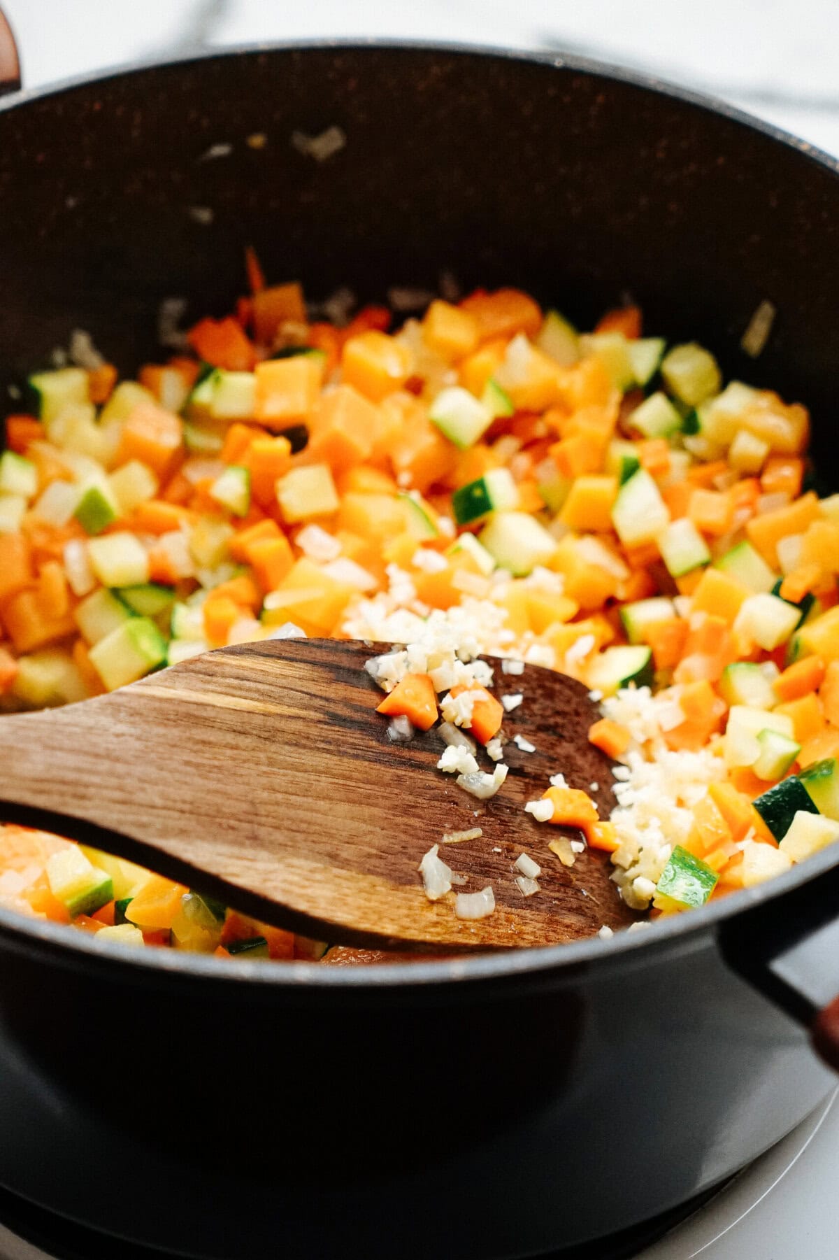 A wooden spatula stirs a mixture of diced vegetables, including squash and zucchini, in a large black pot.