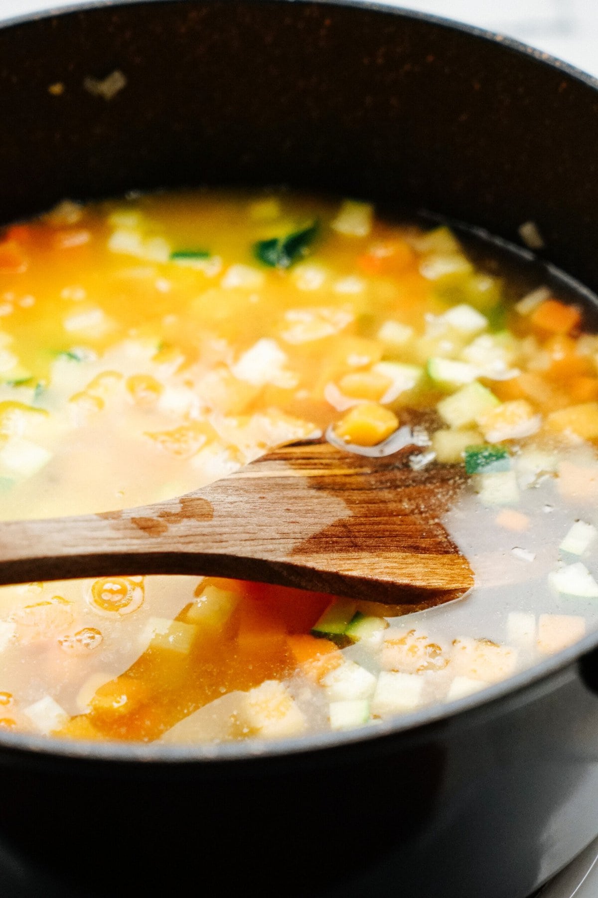 A pot of vegetable soup being stirred with a wooden spoon, showing diced vegetables like carrots, celery, and potatoes in a clear broth.