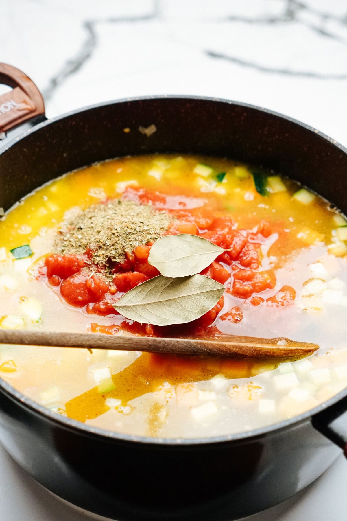 A pot of vegetable soup with diced tomatoes, spices, bay leaves, and a wooden spoon placed across the top.