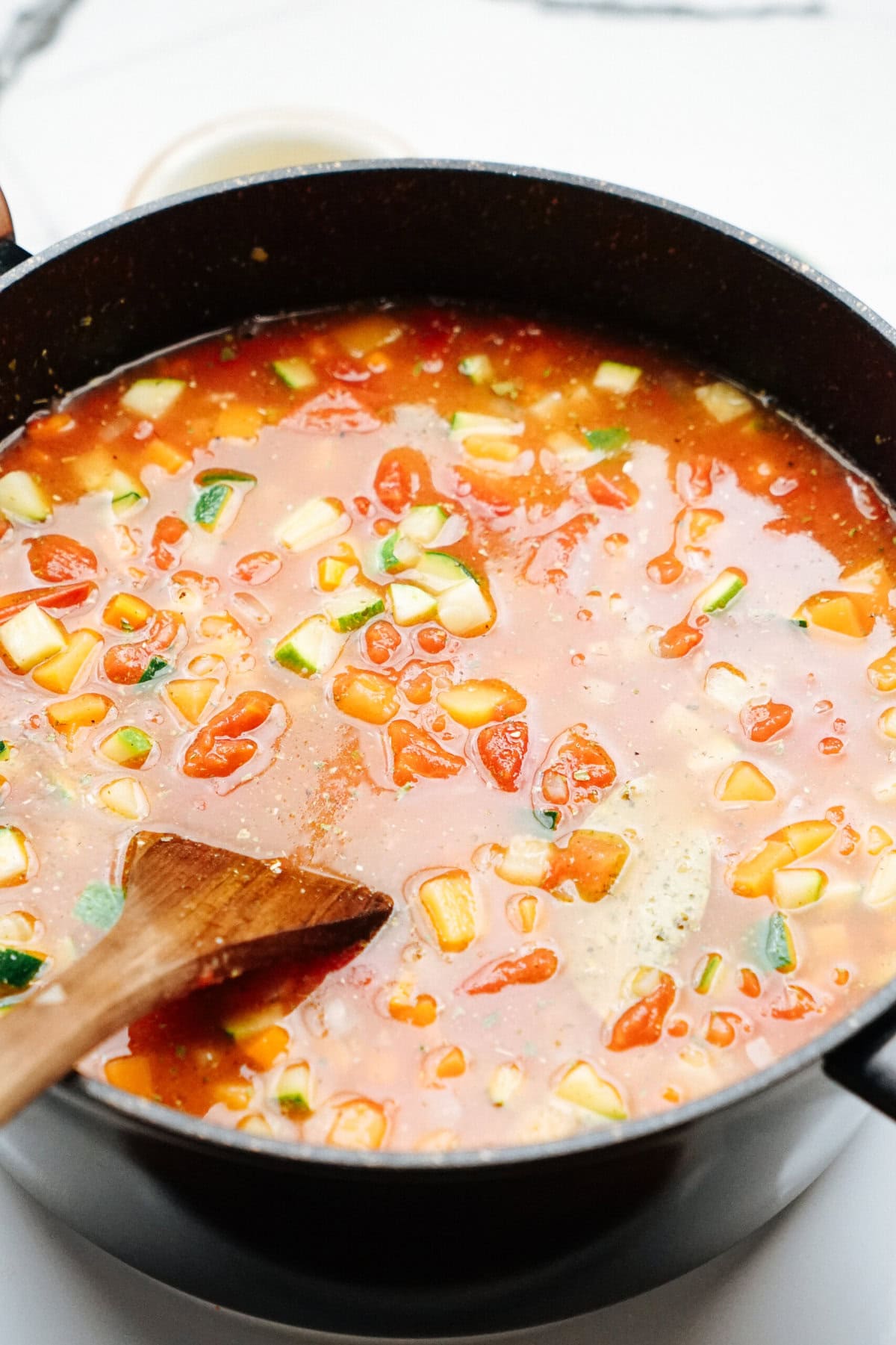 A pot of vegetable soup with chunks of tomato, zucchini, and other vegetables being stirred with a wooden spoon.