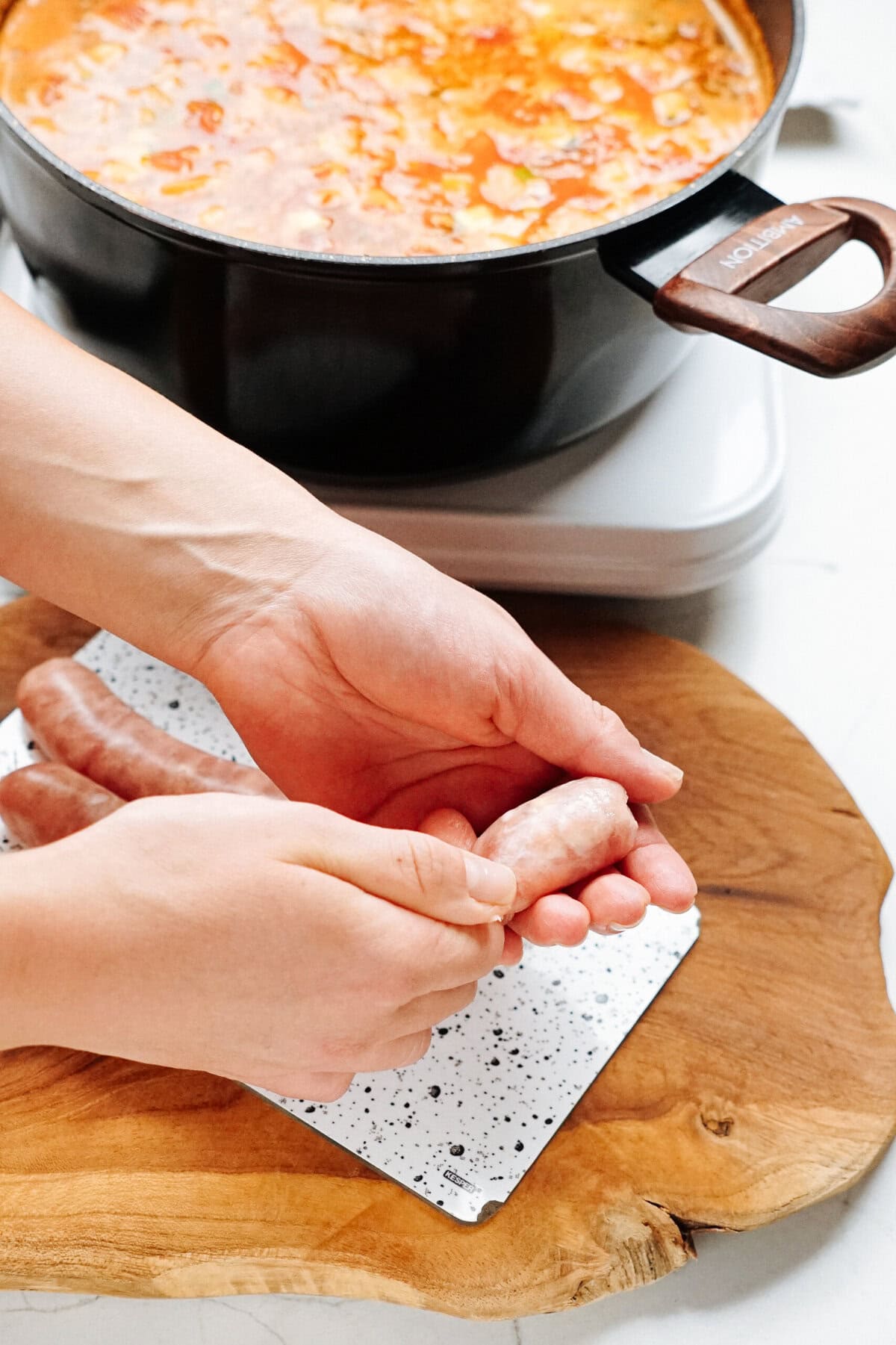 Hands are shaping raw sausages on a wooden board. Behind, a black pot filled with a tomato-based dish is cooking on a stovetop.