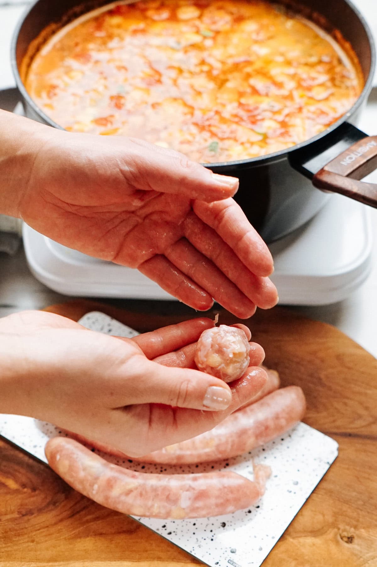 Hands shaping meatballs with uncooked sausages in the background and a pot of soup or stew on the stove.