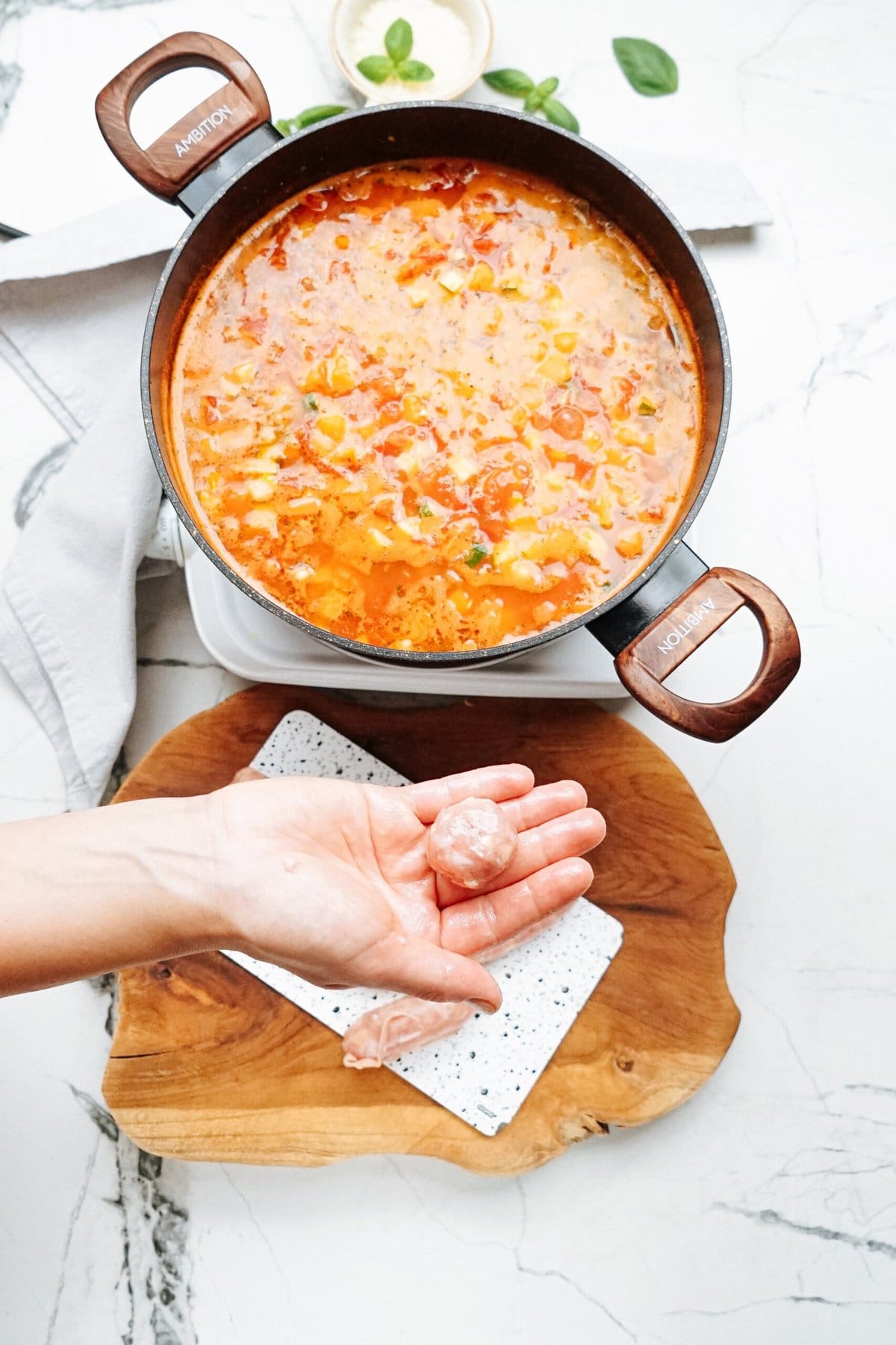 Person holding a small meatball over a pot of soup on a marble countertop with a wooden cutting board nearby.