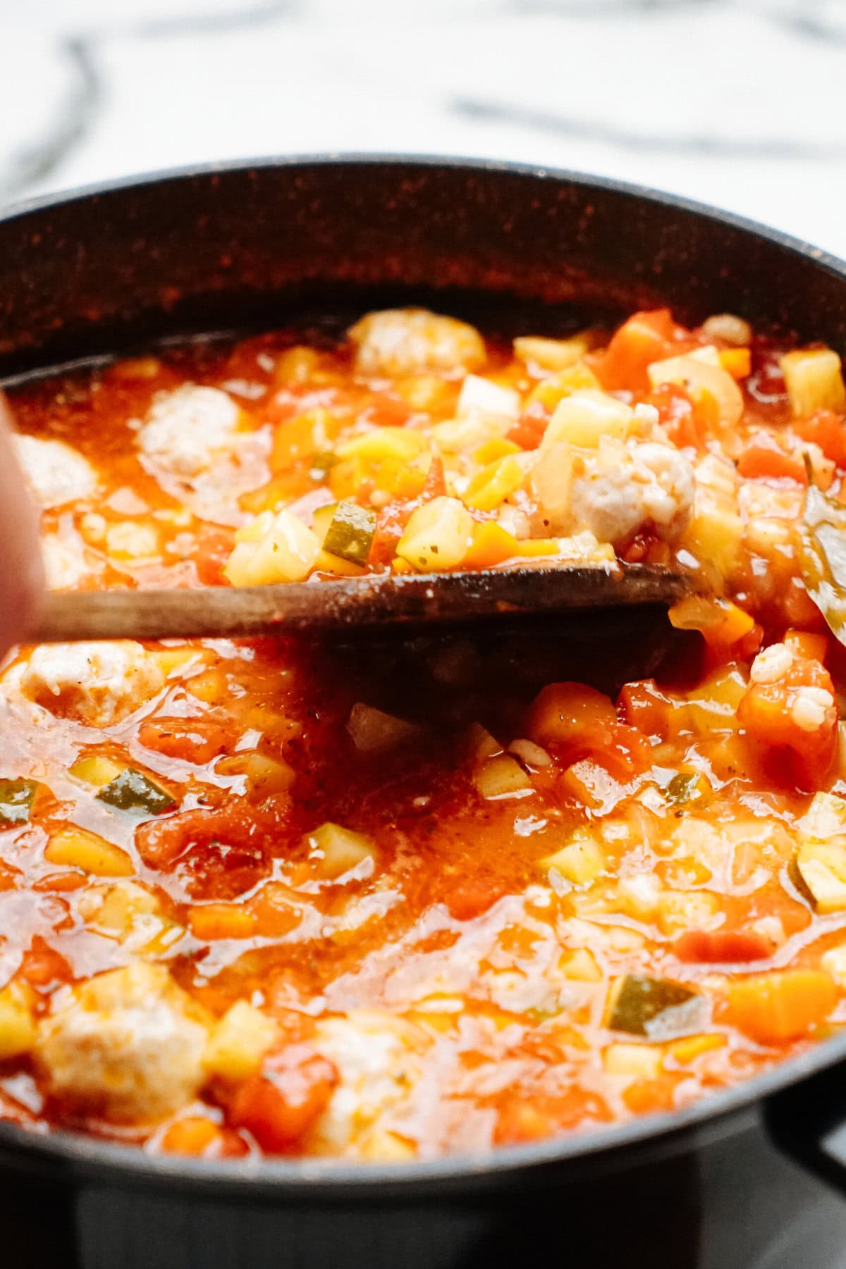 A wooden spoon stirs a pot of chunky vegetable soup with visible pieces of carrots, potatoes, and other vegetables in a tomato-based broth.