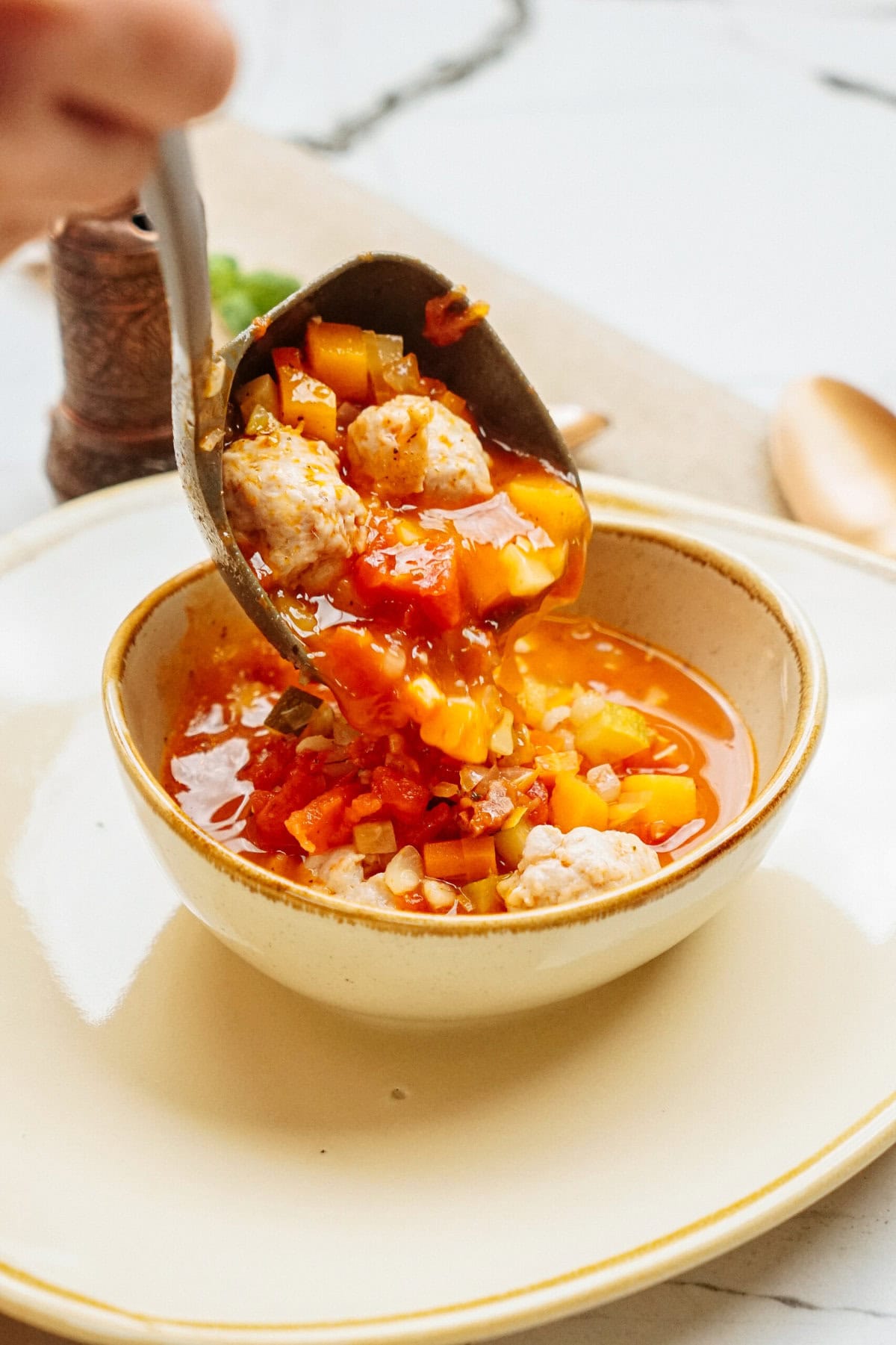 A ladle serves a bowl of chunky chicken meatball soup on a light-colored ceramic plate.