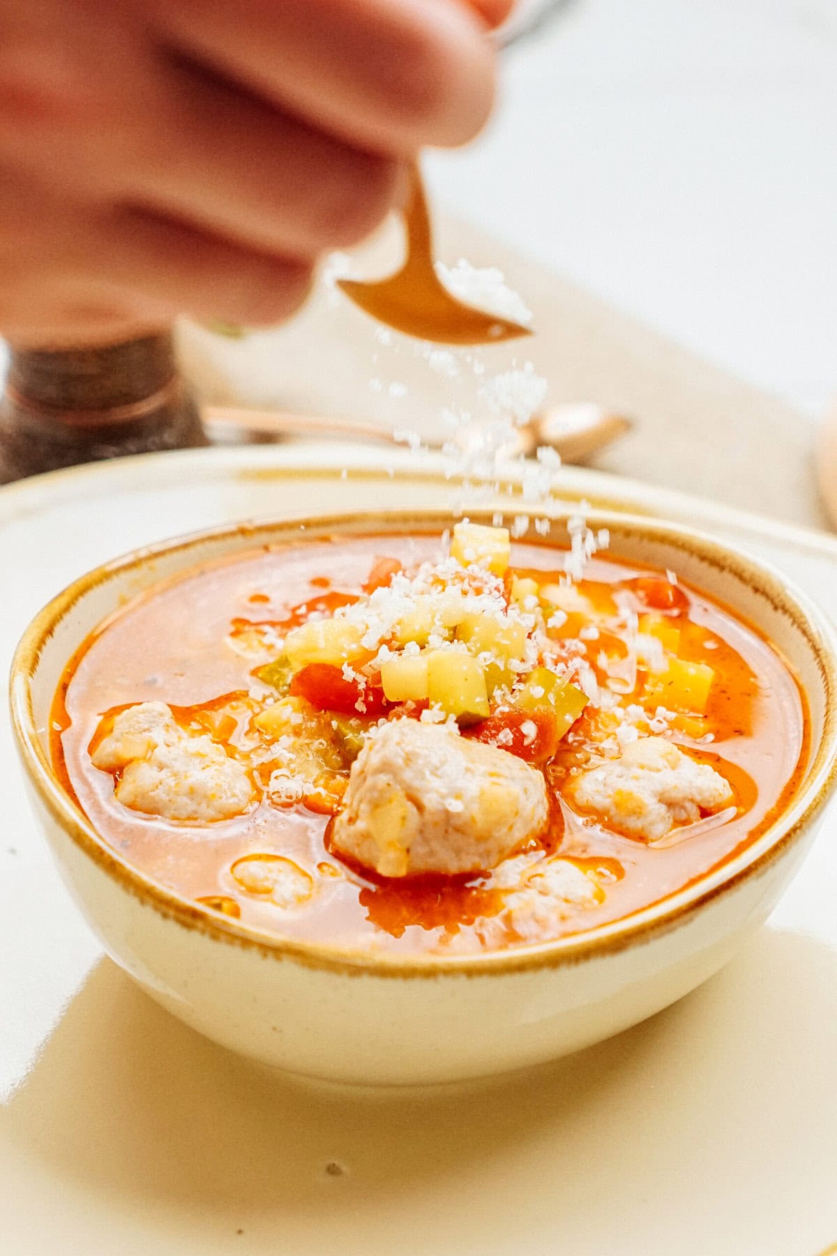 A hand sprinkles grated cheese onto a bowl of soup containing vegetables and meatballs.