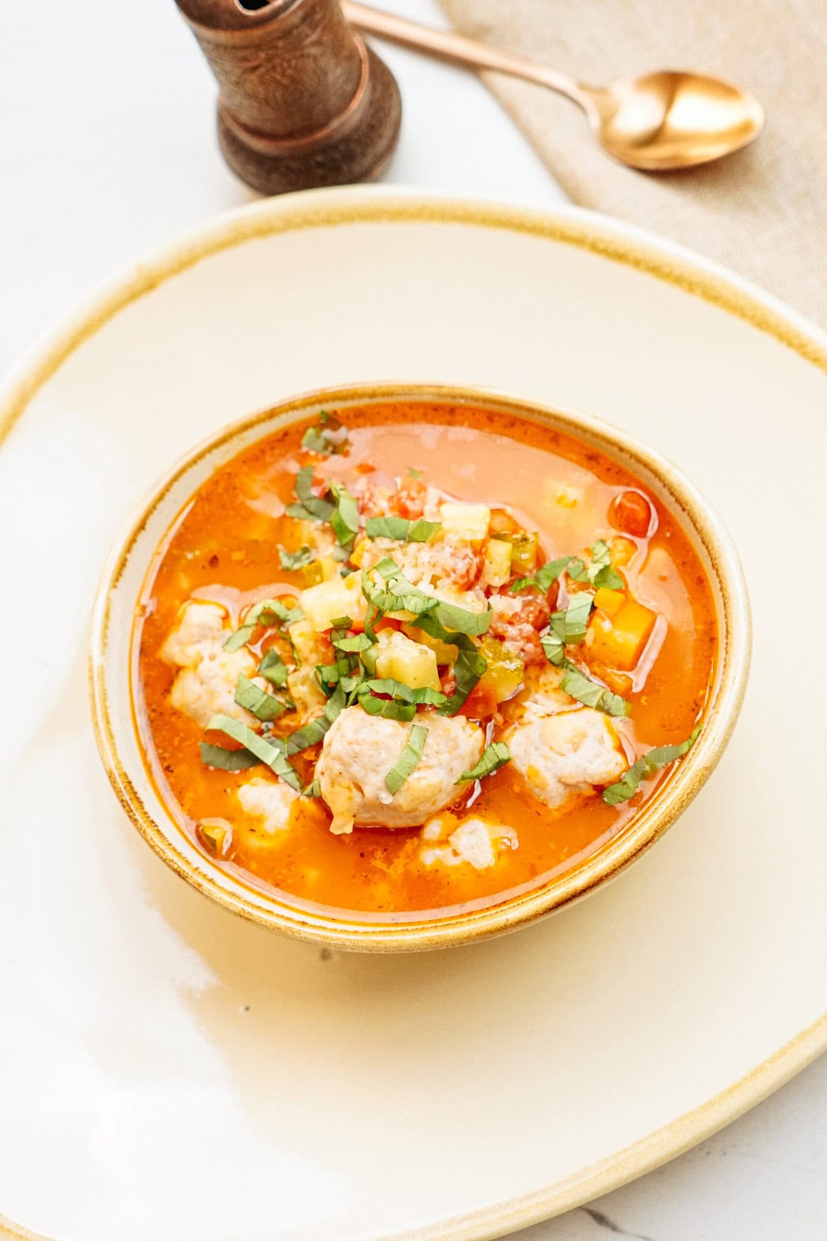 A bowl of red soup with vegetables and meatballs, garnished with chopped herbs, placed on a white plate with a gold spoon and pepper grinder in the background.