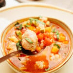 A spoon holding a meatball and vegetables is placed over a bowl of tomato-based soup, garnished with herbs. A spoon and napkin are in the background.