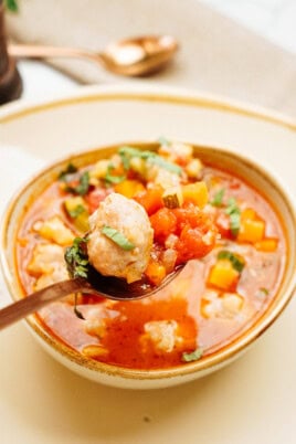 A spoon holding a meatball and vegetables is placed over a bowl of tomato-based soup, garnished with herbs. A spoon and napkin are in the background.