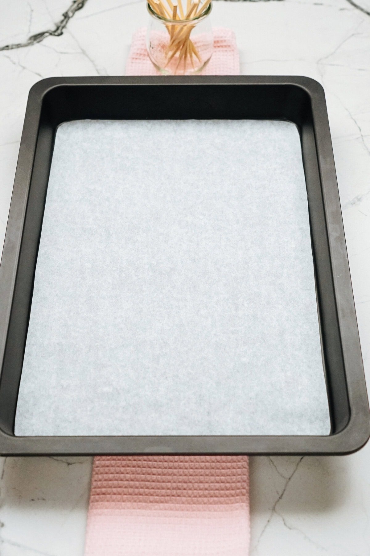 A rectangular baking tray lined with parchment paper rests on a pink textured cloth, ready for gourmet caramel apples. In the background, a small clear jar with wooden skewers awaits its turn in the creation process.