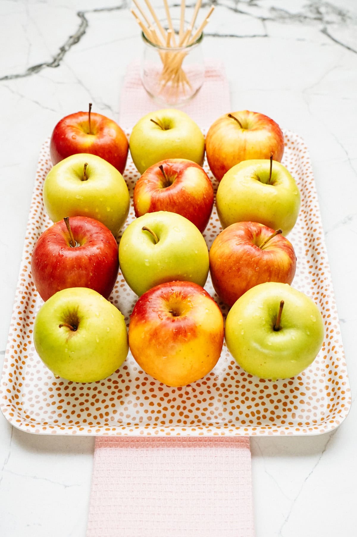 A tray with twelve gourmet caramel apples arranged in rows, featuring a mix of green and red apples. The tray is placed on a light pink cloth with a marble surface in the background.