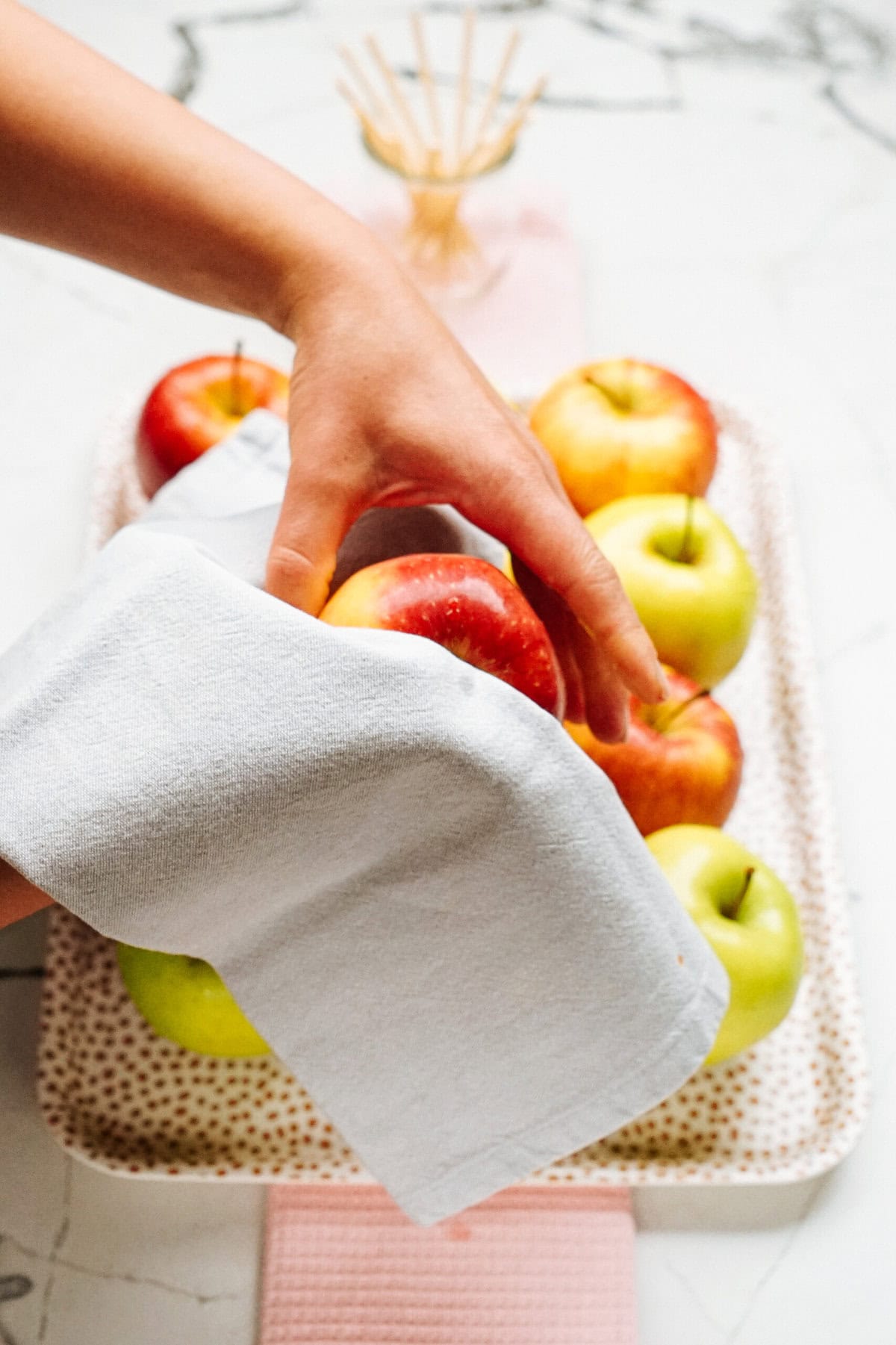 A hand uses a white cloth to polish or clean a red apple among several red and green apples on a dotted tray, preparing them for gourmet caramel apples.