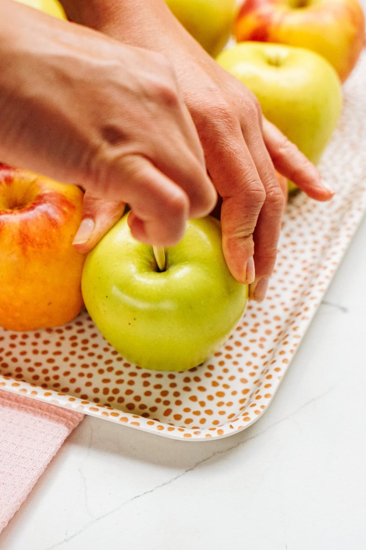 A pair of hands is using an apple corer on a green apple, preparing it for a delicious transformation into gourmet caramel apples. Other apples await their turn on a polka-dotted tray.