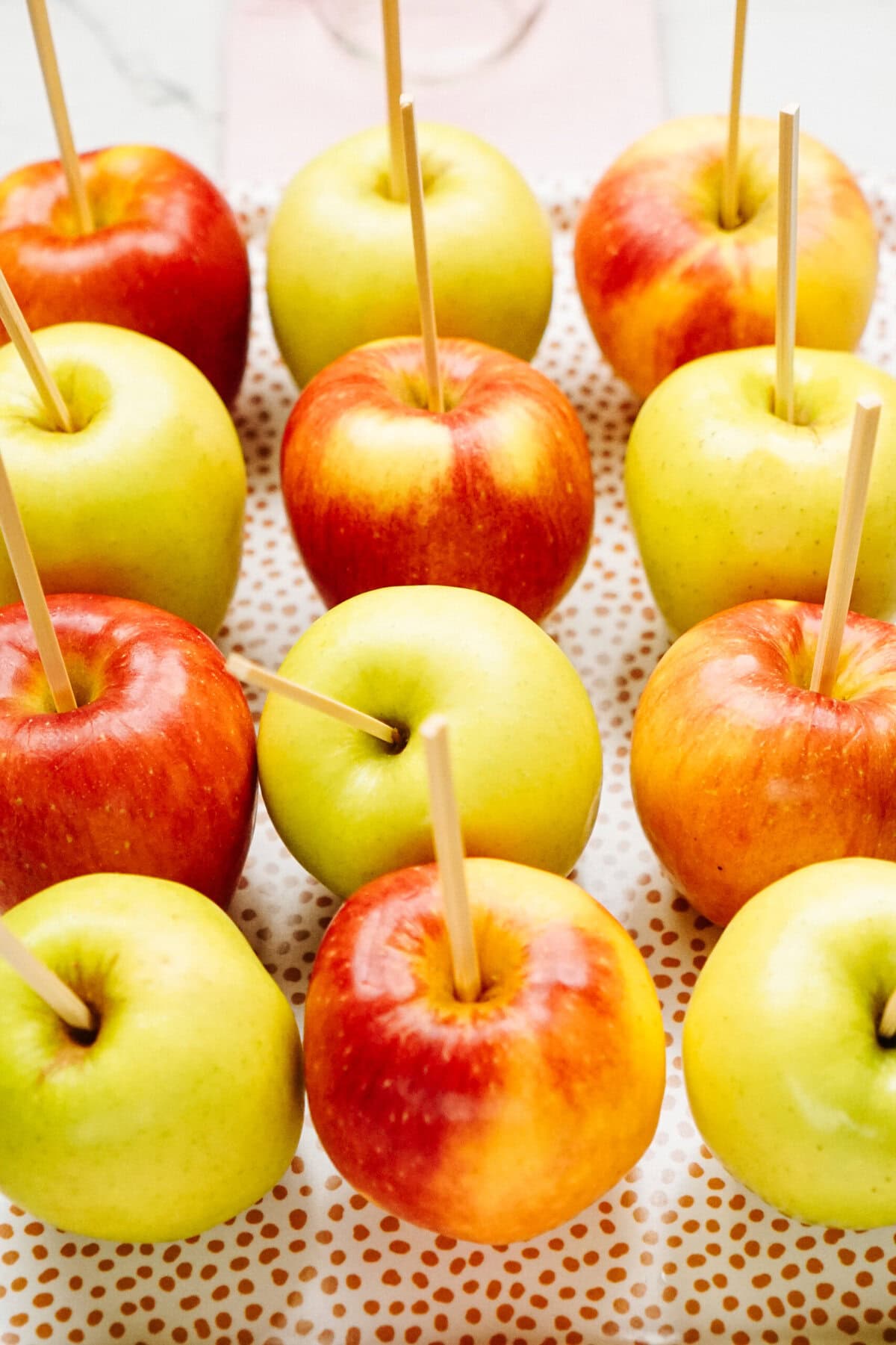 Red and yellow gourmet caramel apples with wooden sticks inserted at the stems, arranged in a grid pattern on a white perforated tray.