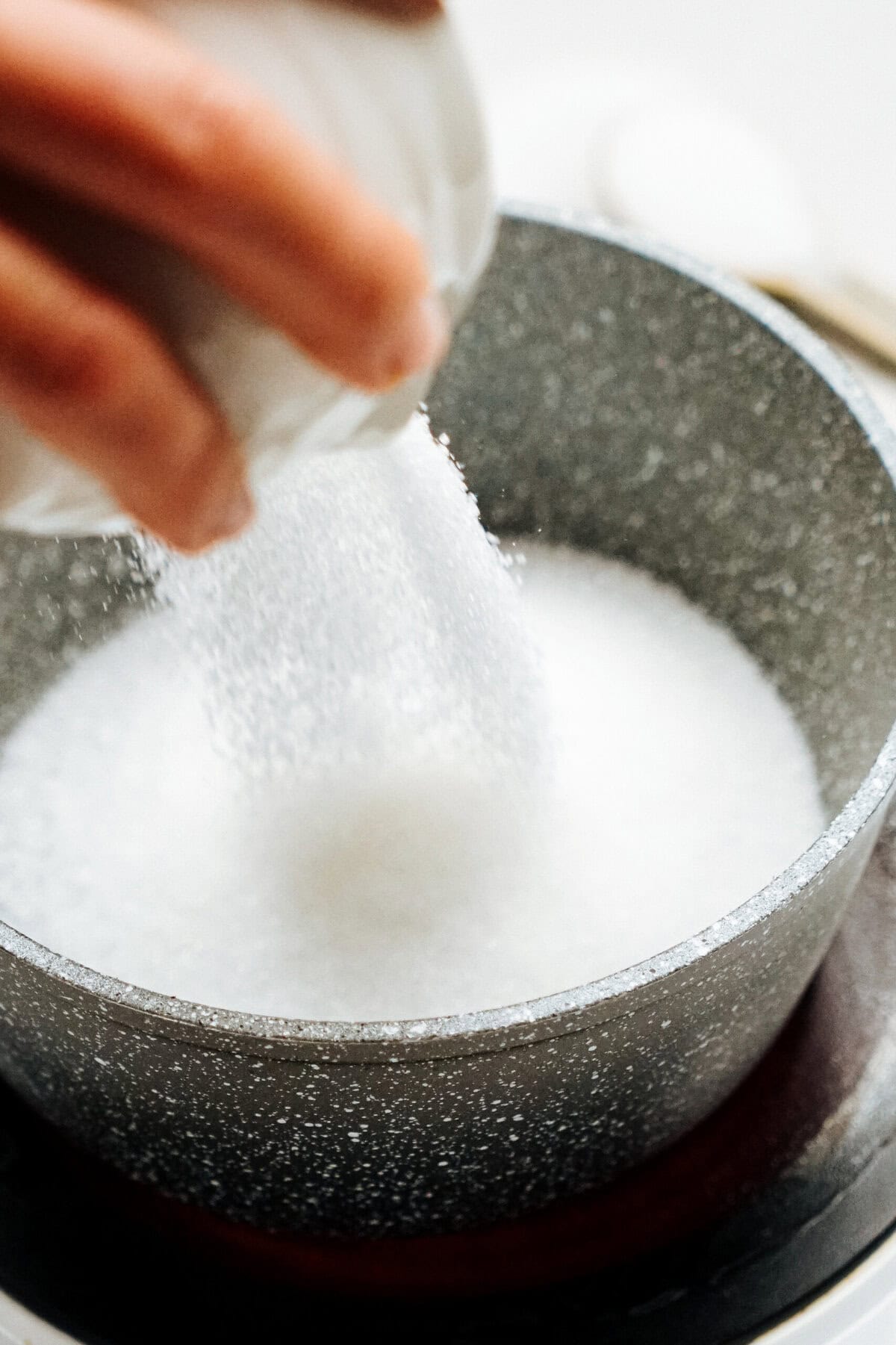 Hand pouring sugar into a gray pot on a stovetop.