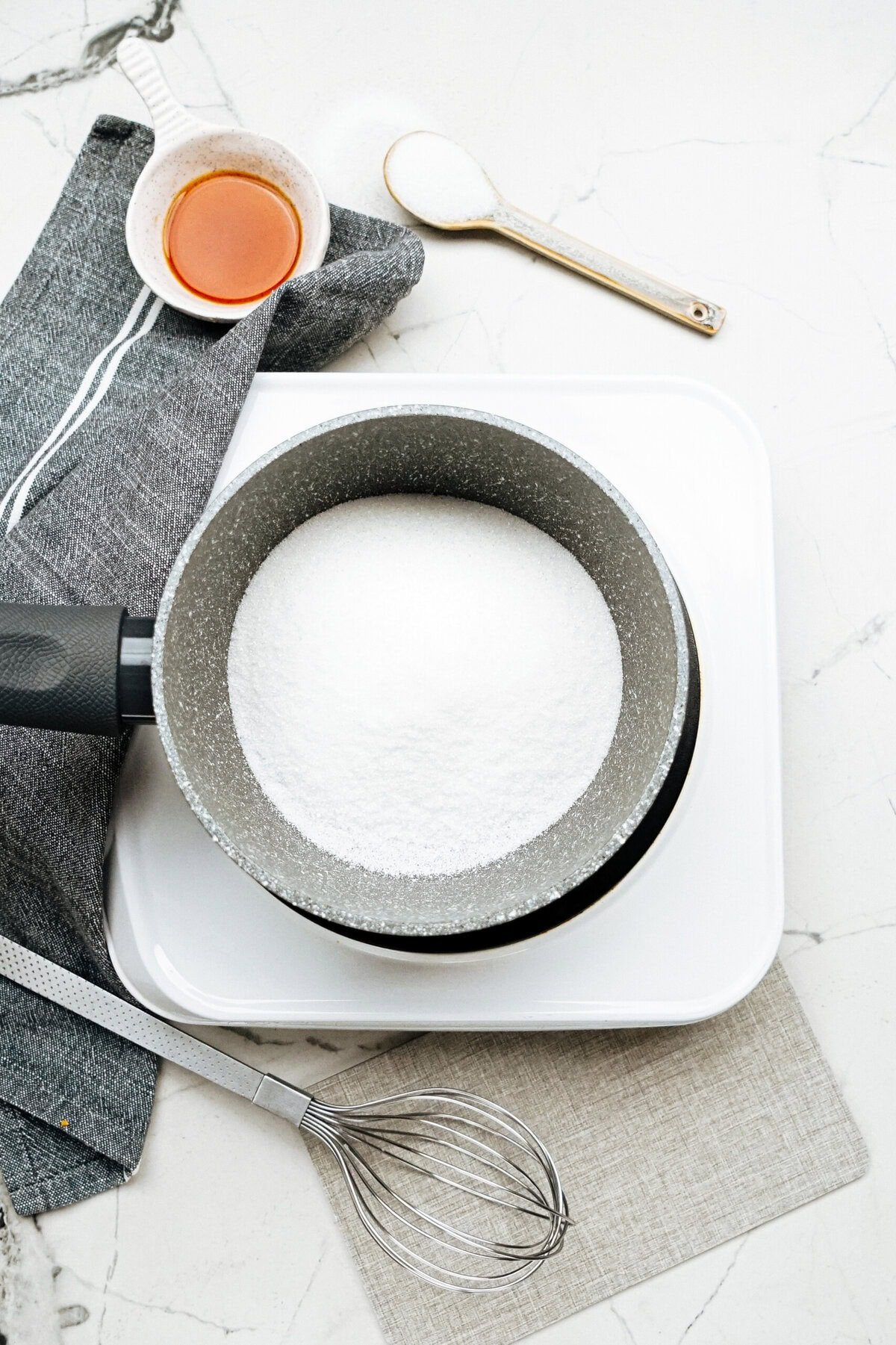 A saucepan filled with white sugar sits on a hot plate, accompanied by a whisk, a cloth, a small bowl of liquid, and a spoon on a marble countertop—perfect for making gourmet caramel apples.