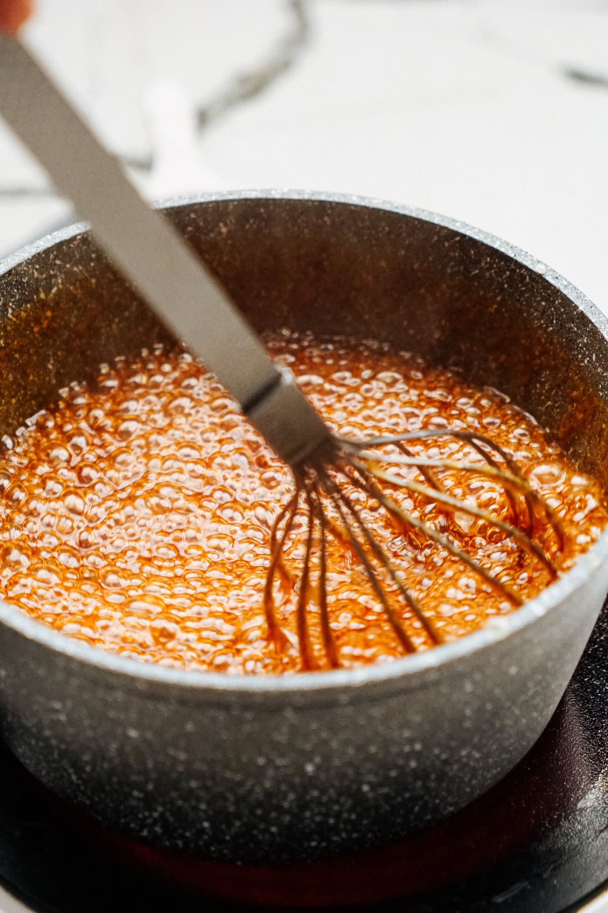 A hand holding a whisk stirs a bubbling caramel mixture in a dark gray saucepan on a white stovetop, preparing it for gourmet caramel apples.