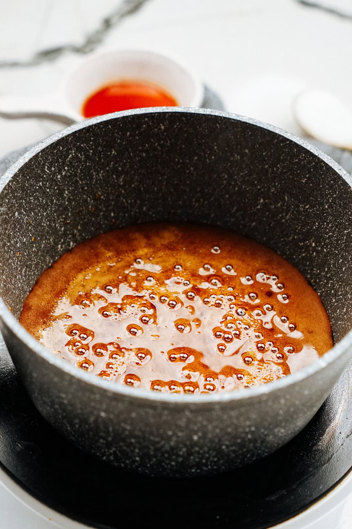 A pot on a stovetop contains bubbling caramel mixture, with a small bowl of caramel sauce in the background.