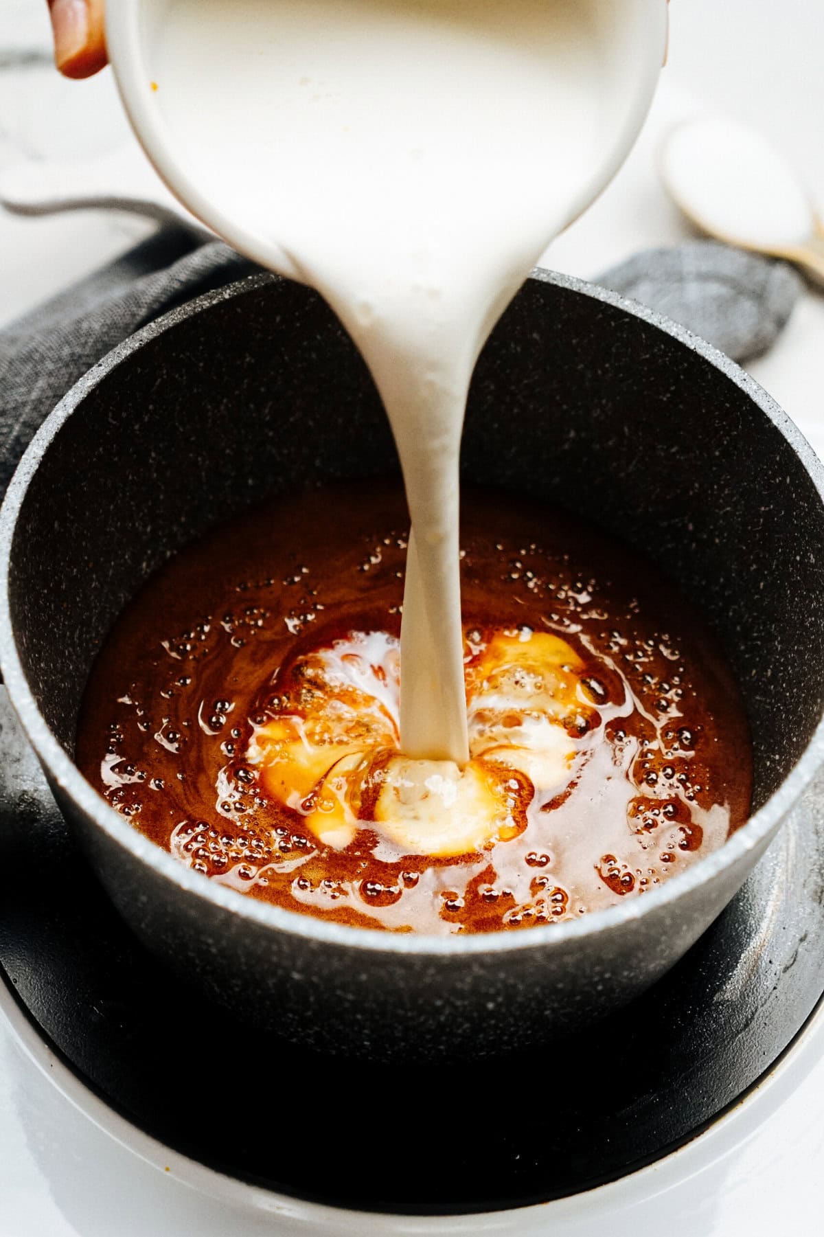 A stream of cream is being poured into a dark saucepan containing a reddish-brown mixture, likely gourmet caramel or sauce for making delectable caramel apples, on a stovetop.