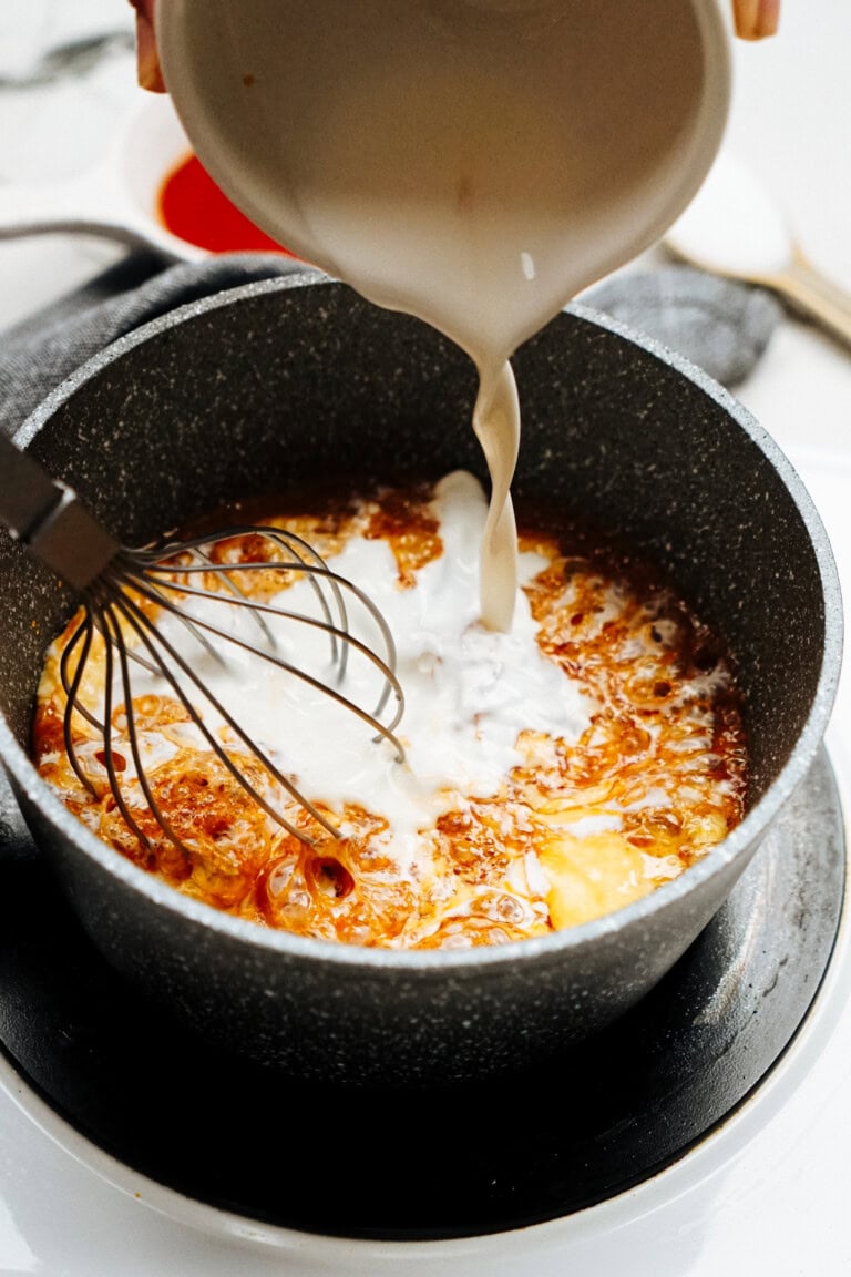 A close-up view of a creamy liquid being poured into a pot of caramel sauce on a stove, with a whisk placed inside the pot.