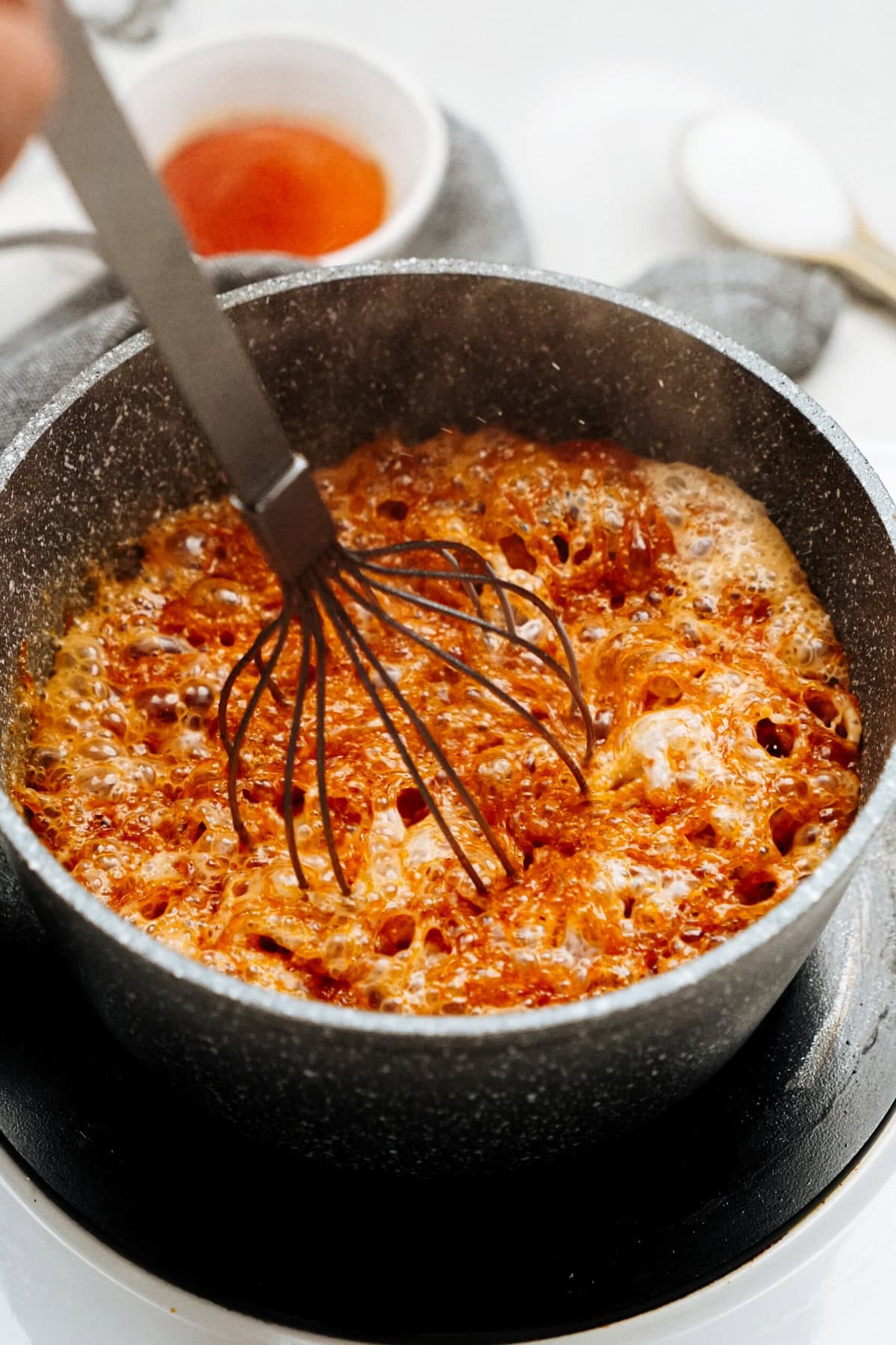 A whisk stirs a bubbling caramel mixture in a black pot on the stovetop, with a cup of gourmet caramel sauce in the background—perfect for creating decadent caramel apples.