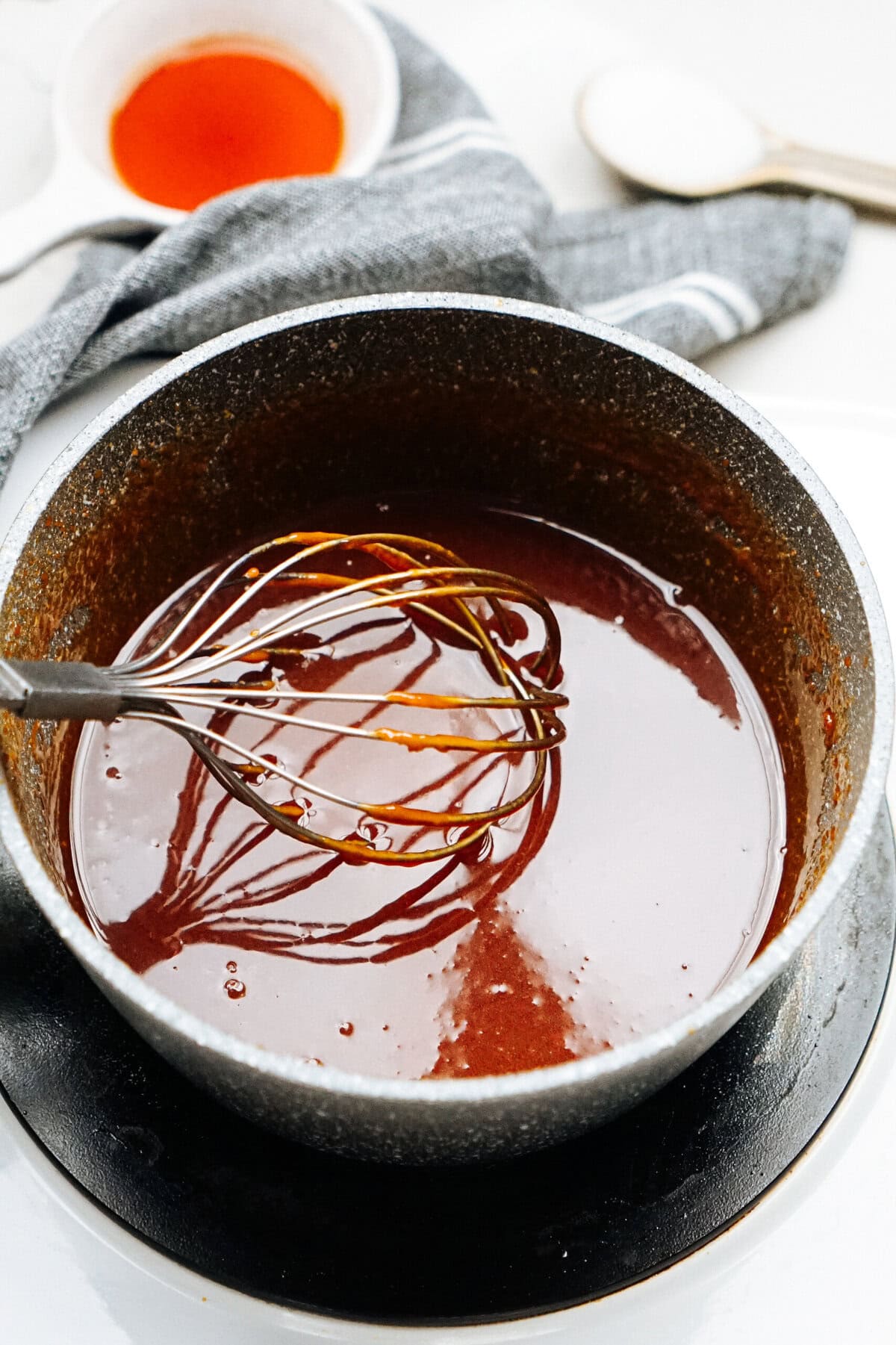 A saucepan filled with dark sauce, a whisk resting inside. A tea towel, a small cup and a spoon are in the background.