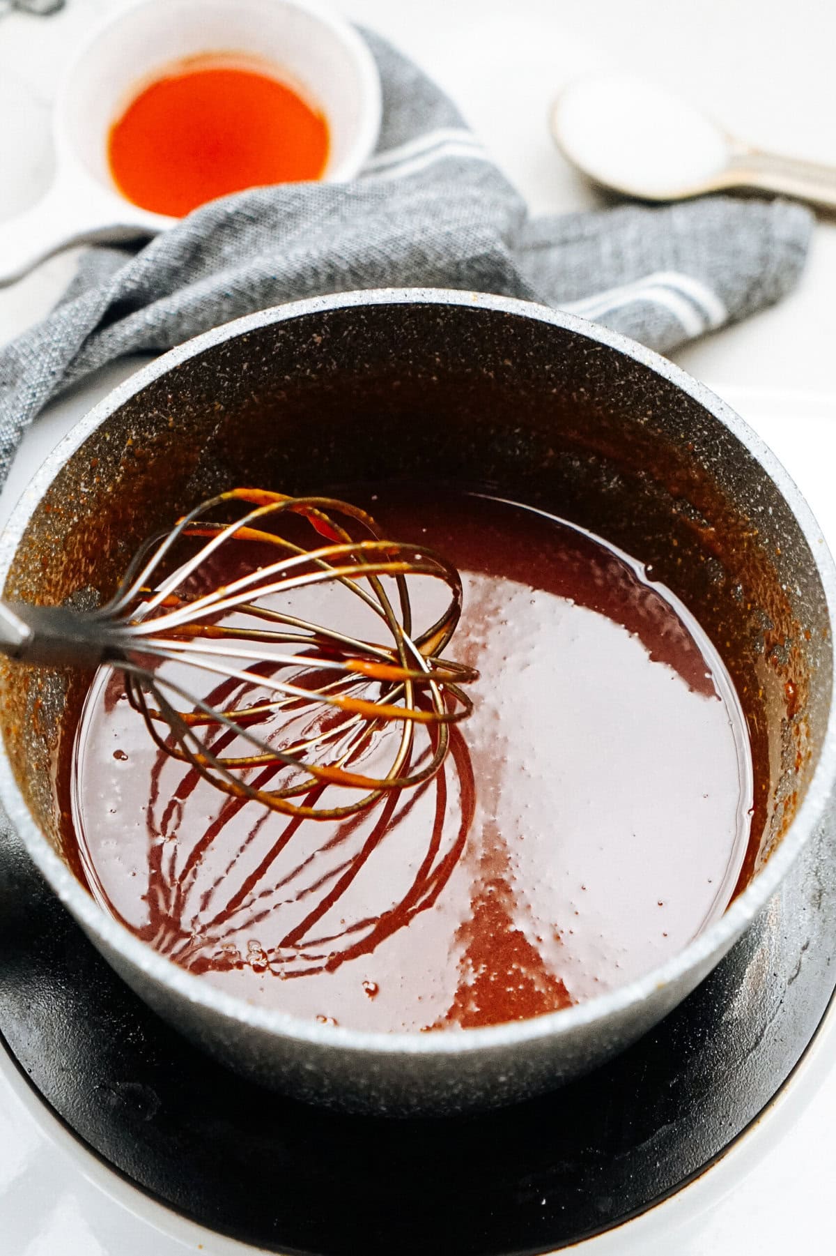 A whisk stirs a dark, thick sauce in a pot, reminiscent of the rich coating on gourmet caramel apples. In the background, a bowl with a similar sauce, alongside a cloth and spoon, completes the culinary scene.