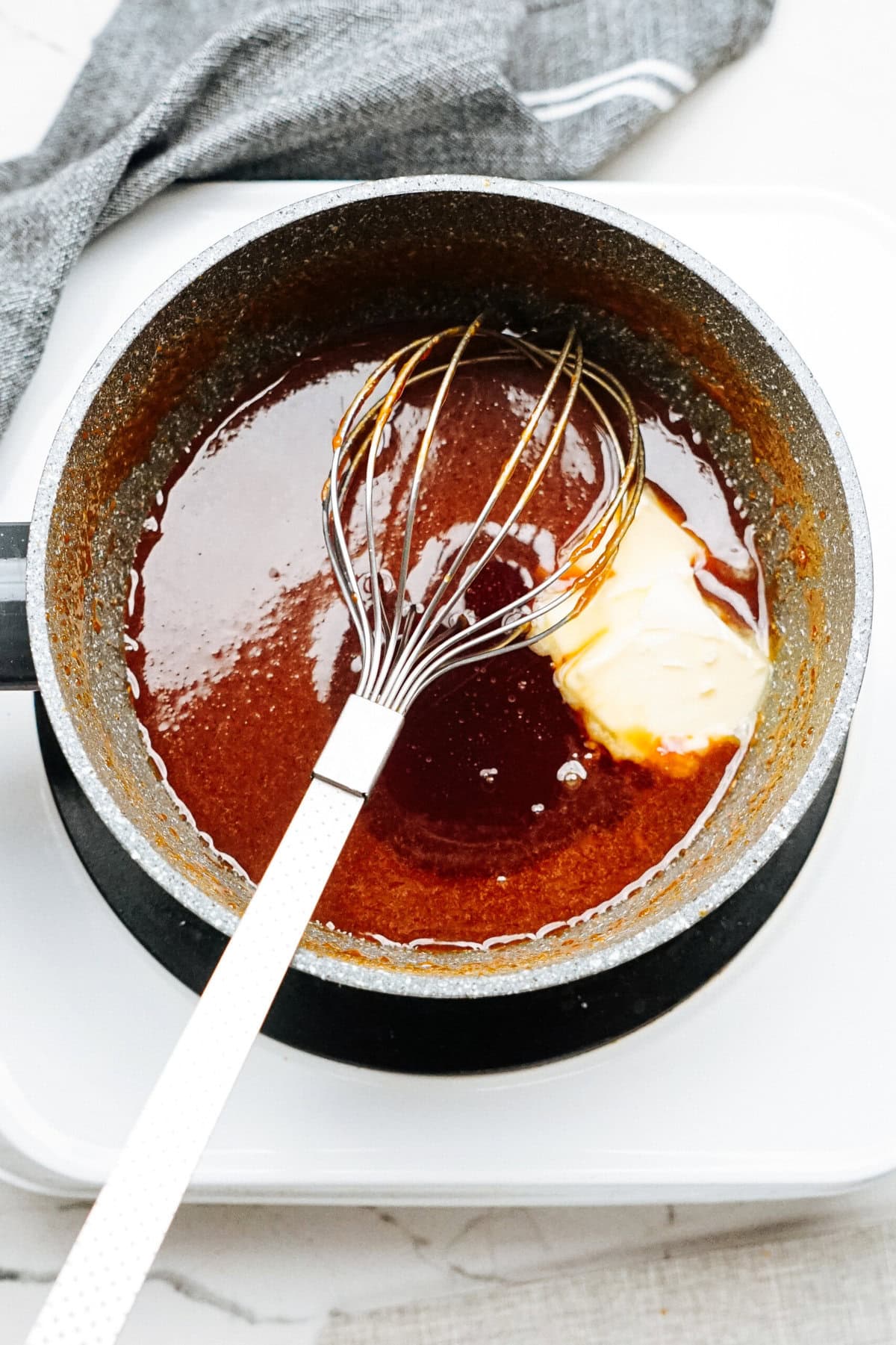 A whisk rests inside a saucepan filled with dark sauce, reminiscent of the rich coatings used for gourmet caramel apples, with a small amount of butter melting into it.