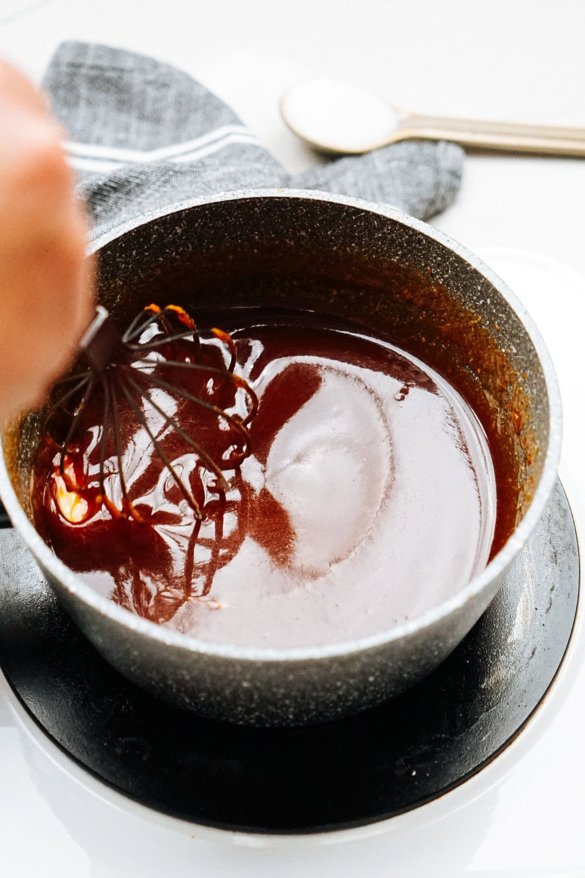 A close-up of a person stirring a thick, dark sauce with a whisk in a small saucepan placed on a black saucer.