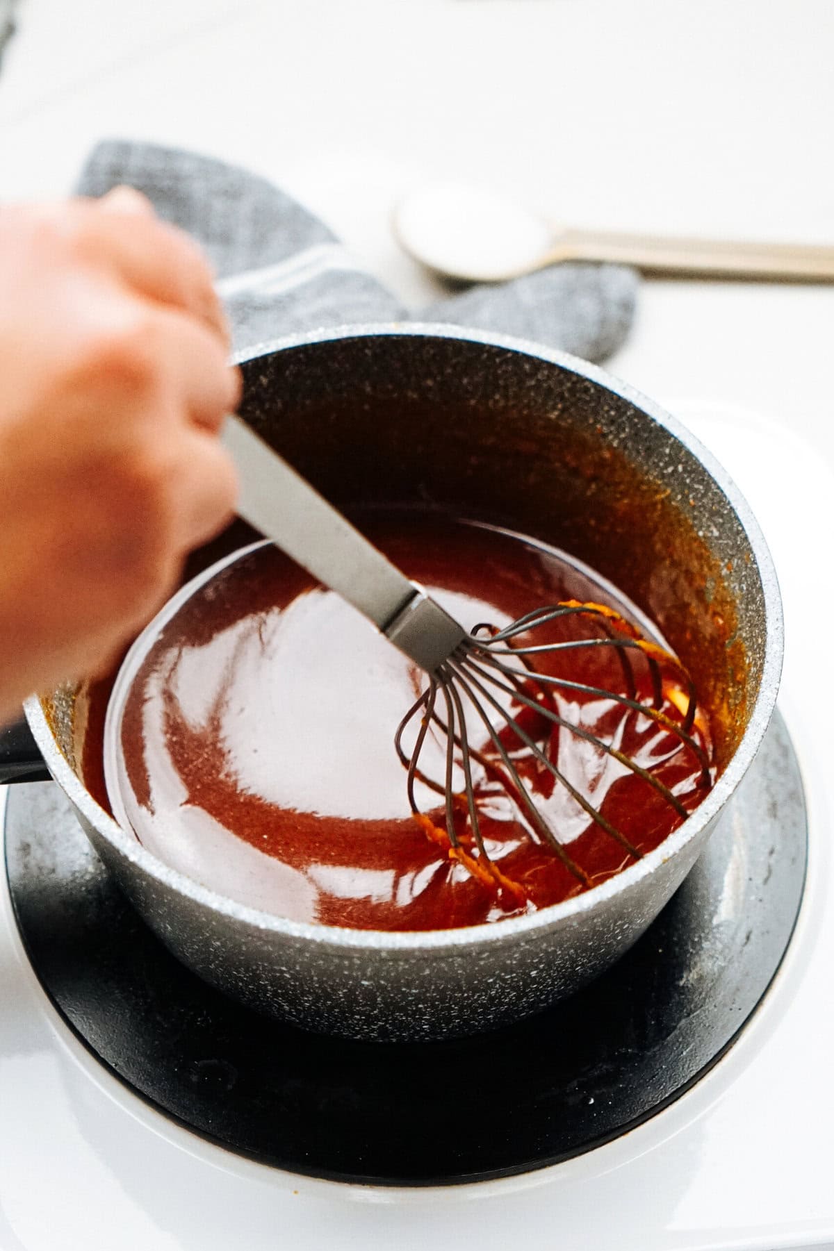 A hand is expertly whisking a dark sauce in a pot on the stove, preparing the perfect drizzle for gourmet caramel apples.