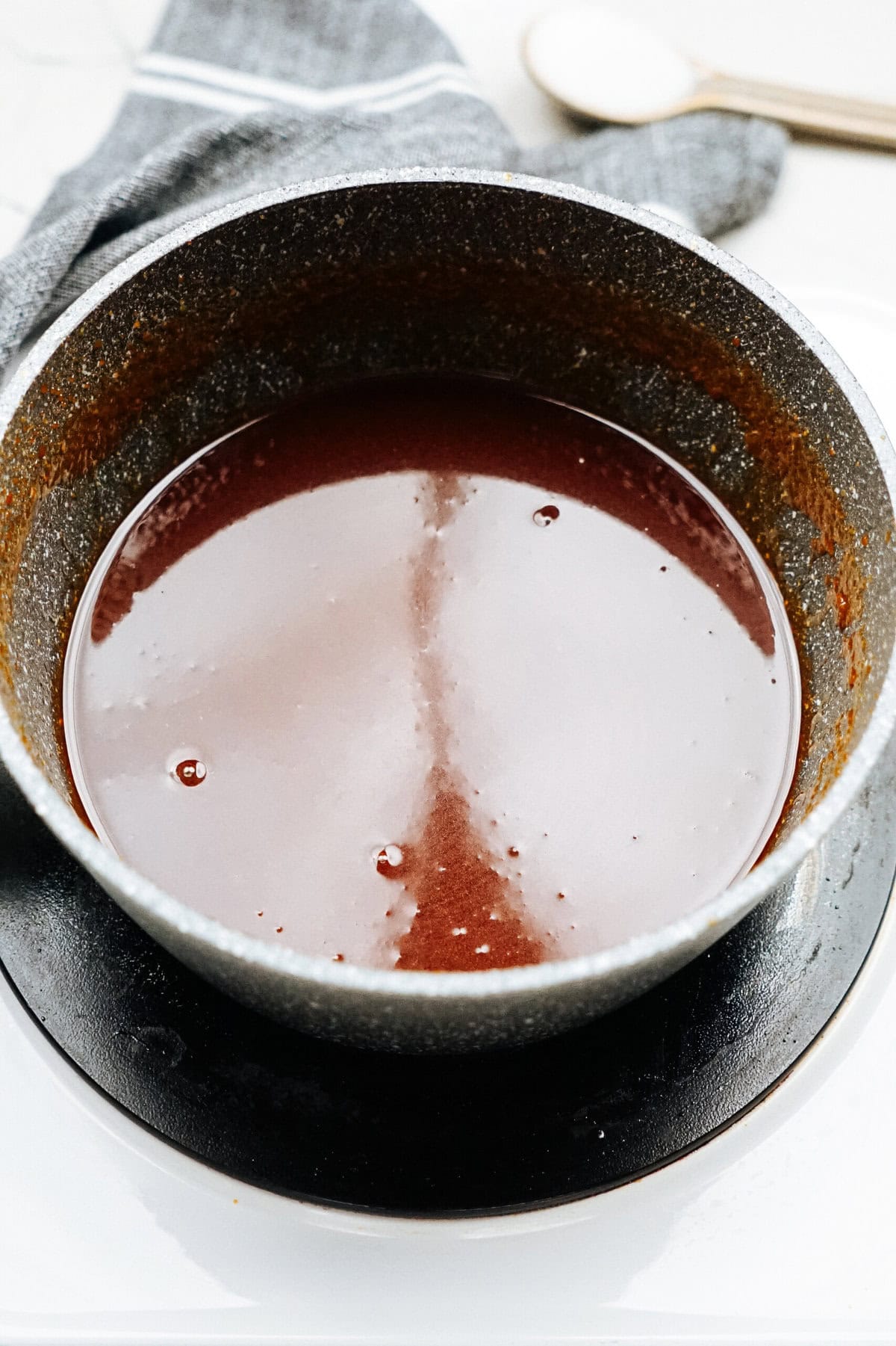 A metal pot filled with a dark red liquid, possibly a sauce or broth, sits on a black plate atop a gray cloth. A wooden spoon rests in the background.