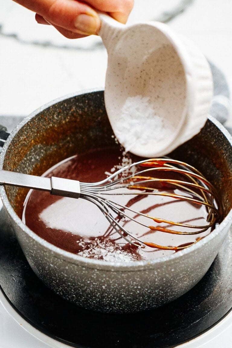 A hand is pouring salt from a small white bowl into a saucepan with chocolate mixture being whisked.