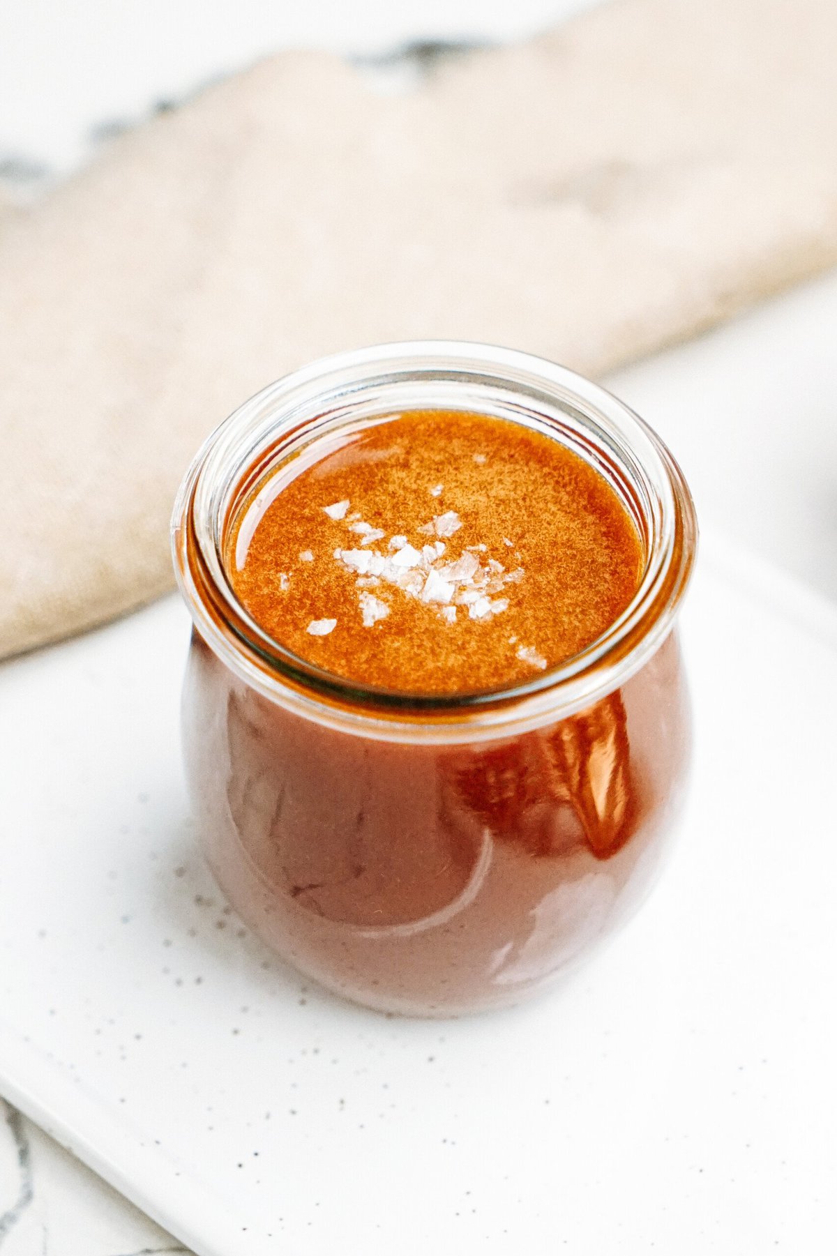 A small glass jar filled with a thick brown liquid topped with coarse sea salt, placed on a white speckled tray with a blurred beige cloth in the background.